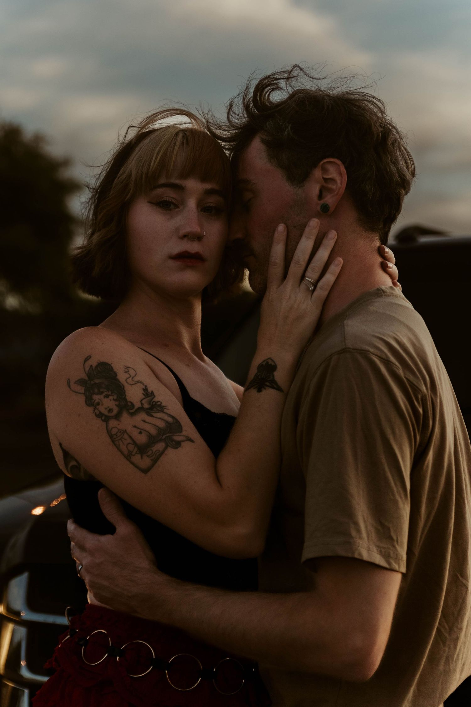 Couple embracing intimately outdoors at dusk, woman with tattoos in black top, man in olive tee whispering in her ear.