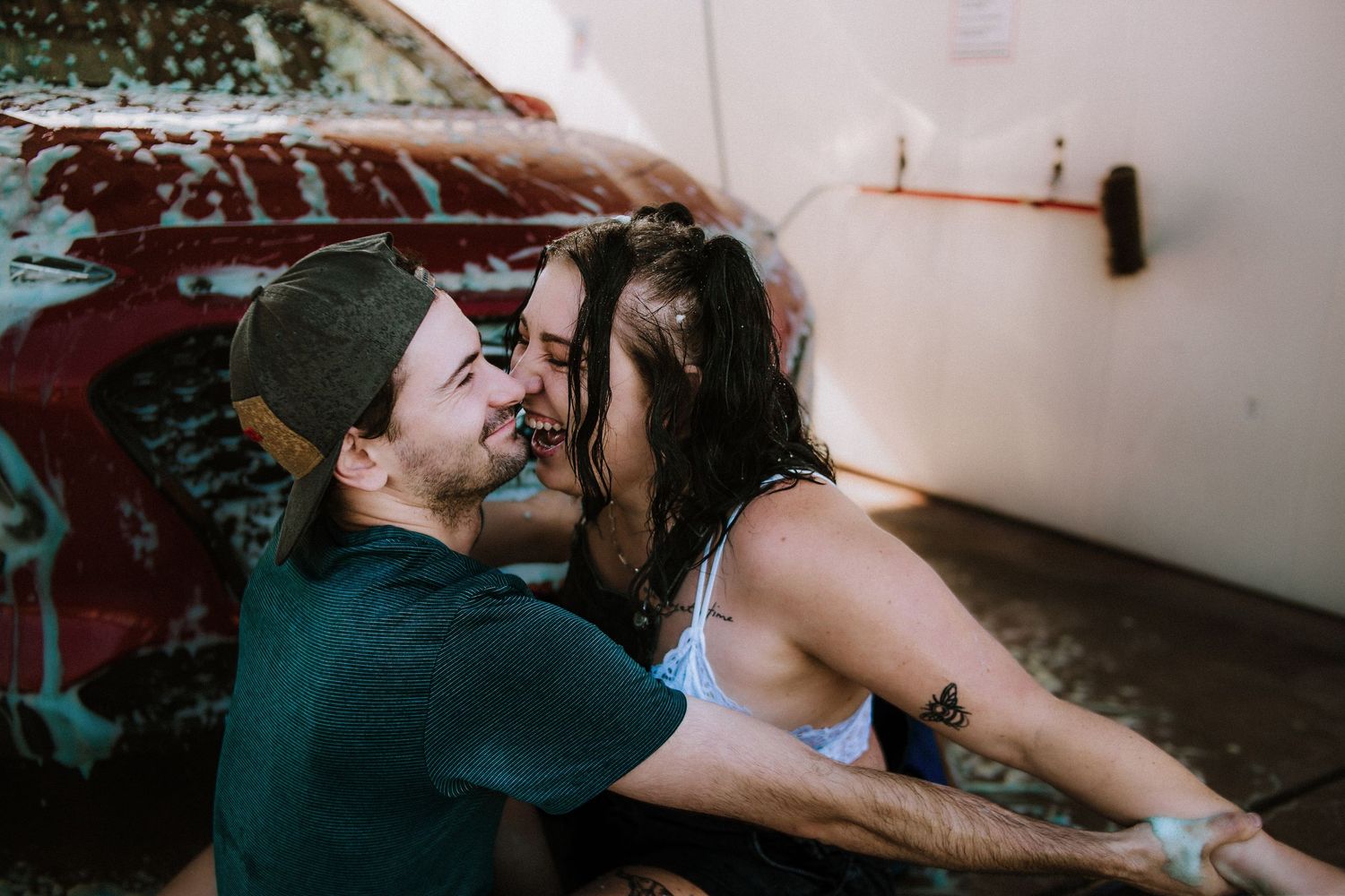 A couple laughs and leans in for a kiss while washing a red car covered in soap suds.