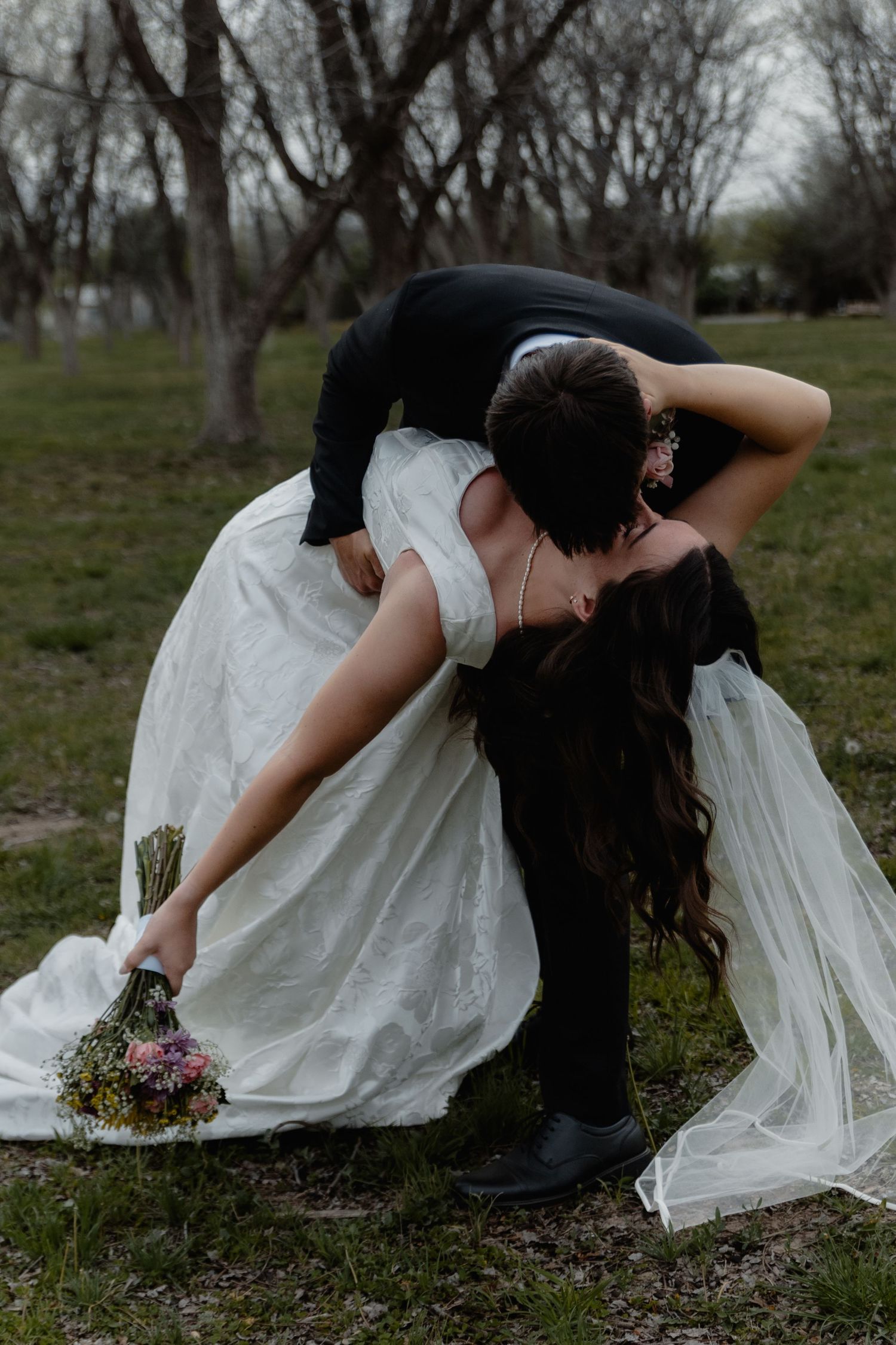 Dramatic wedding photo of couple in a dip pose with bride in flowing white dress holding bouquet against moody background.