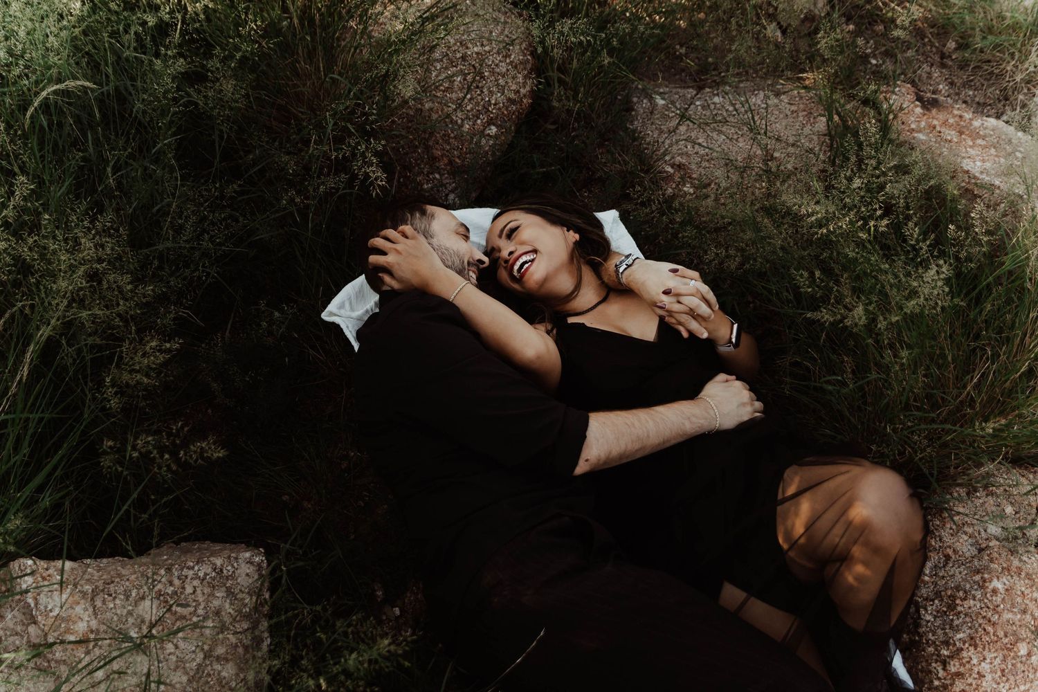 Couple lying on rocks surrounded by greenery, laughing and embracing in a romantic outdoor engagement photo.