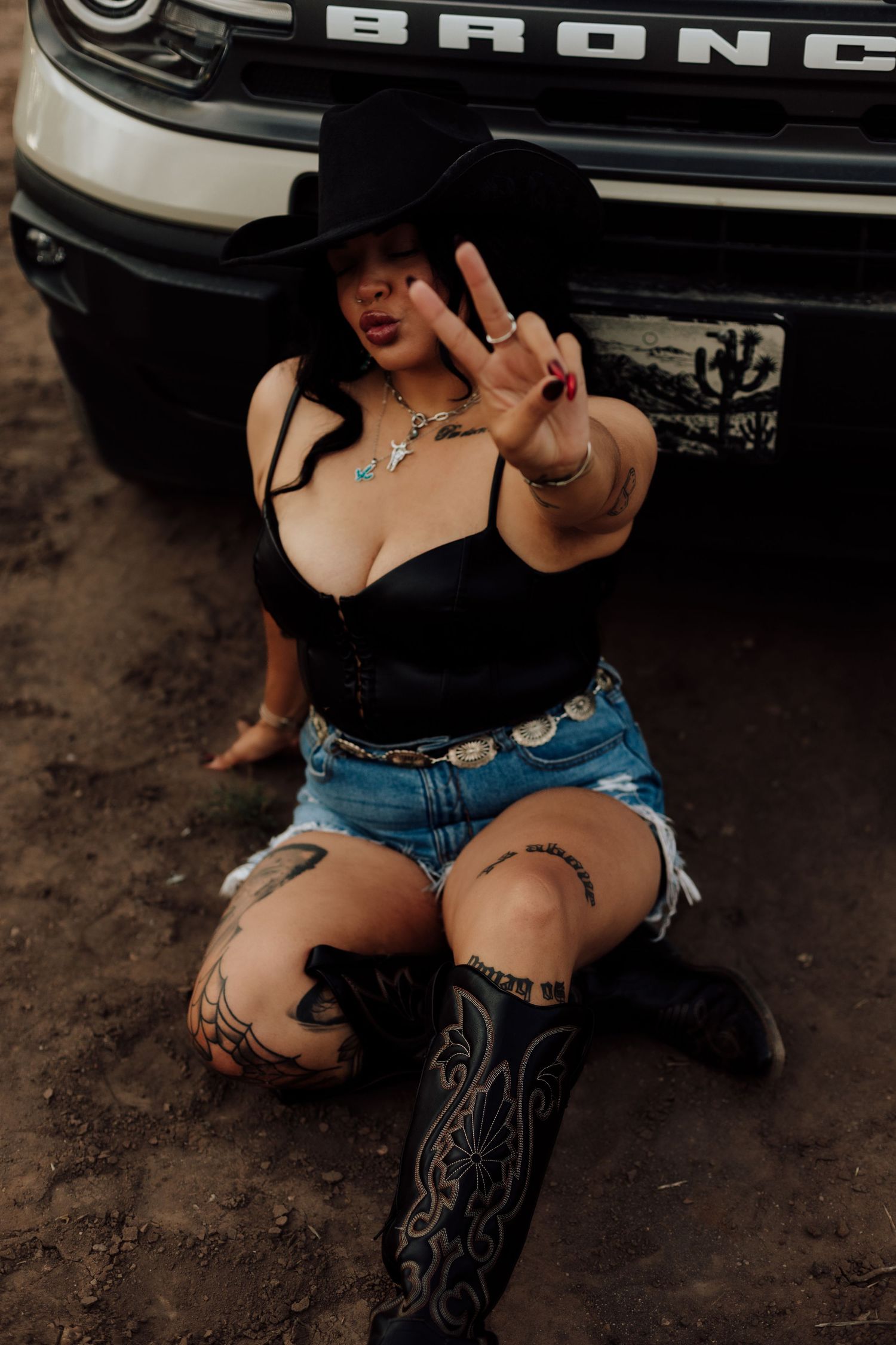 Tattooed woman in black cowboy hat, crop top, denim shorts, and boots sits beside a Ford Bronco flashing a peace sign.