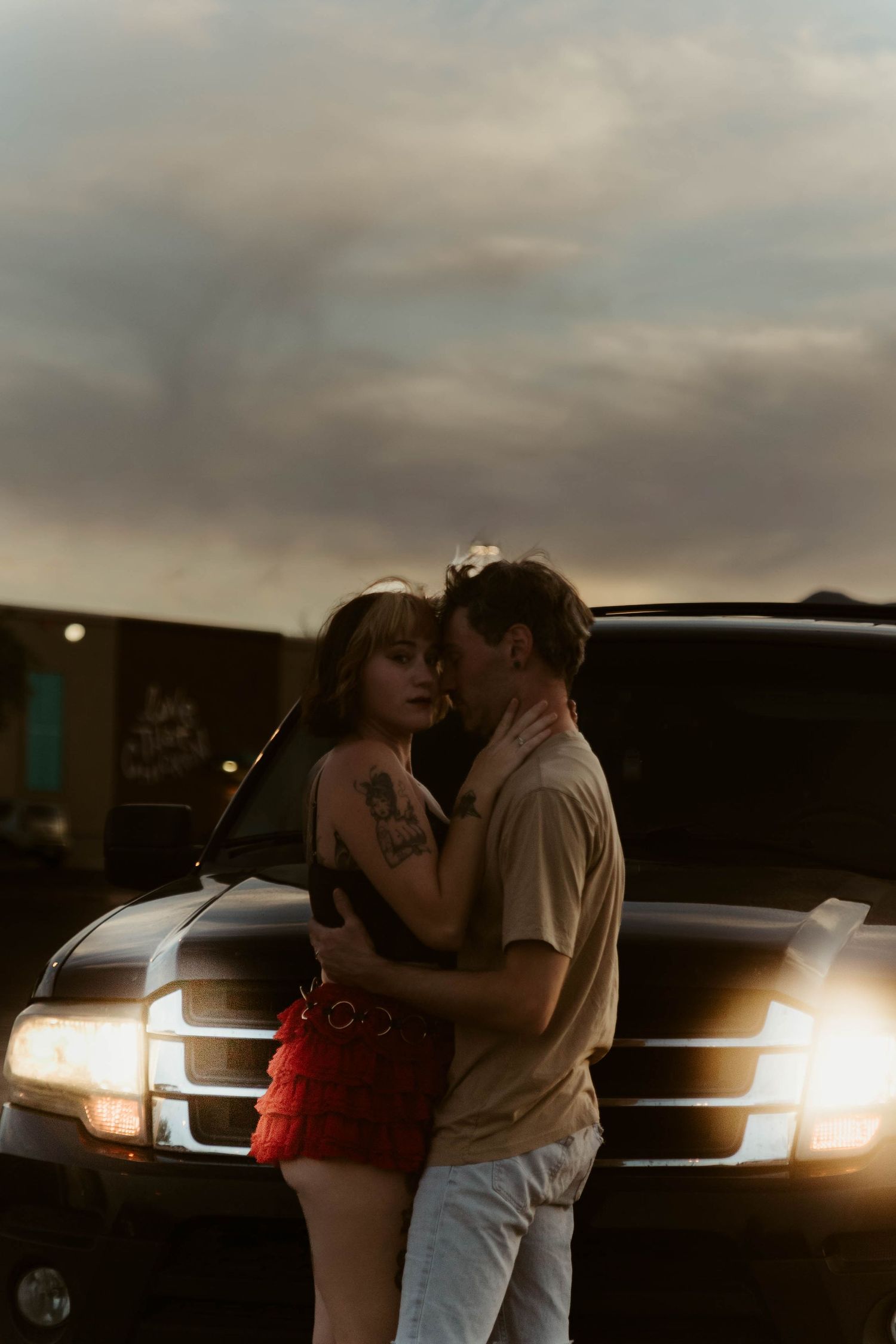 Couple embracing romantically in front of glowing SUV headlights at dusk, woman with tattoos in red skirt.