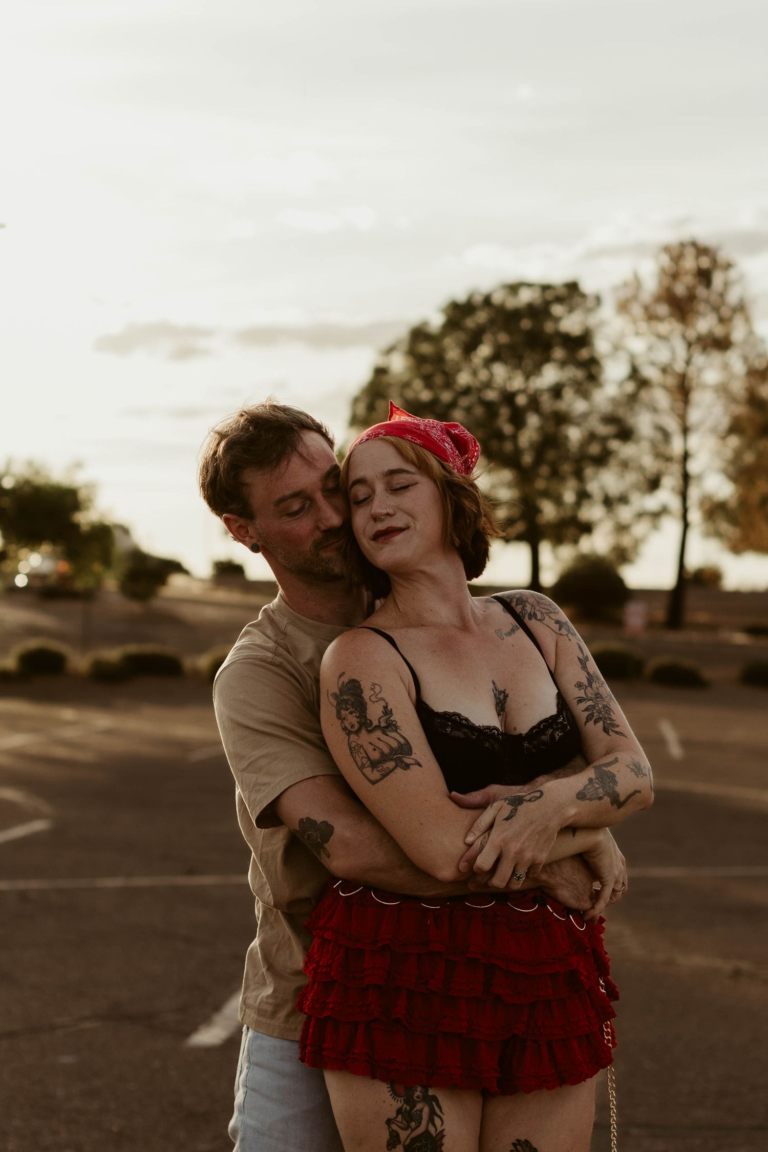 Tattooed couple embracing romantically in a parking lot at golden hour, woman in red skirt and black bralette.