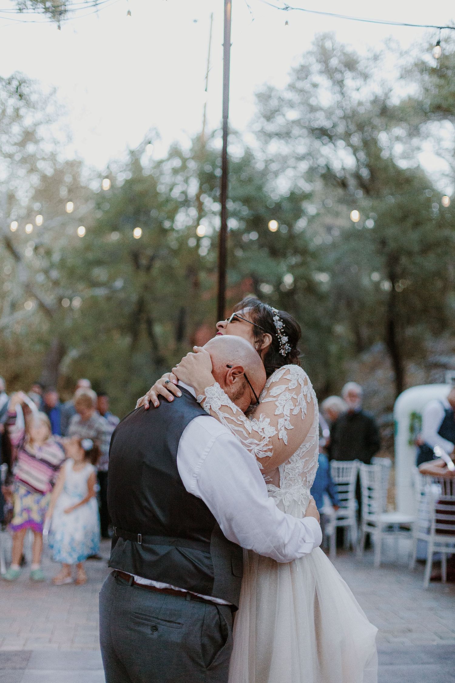 Bride and groom share an emotional first dance embrace outdoors at a romantic evening wedding reception with string lights.
