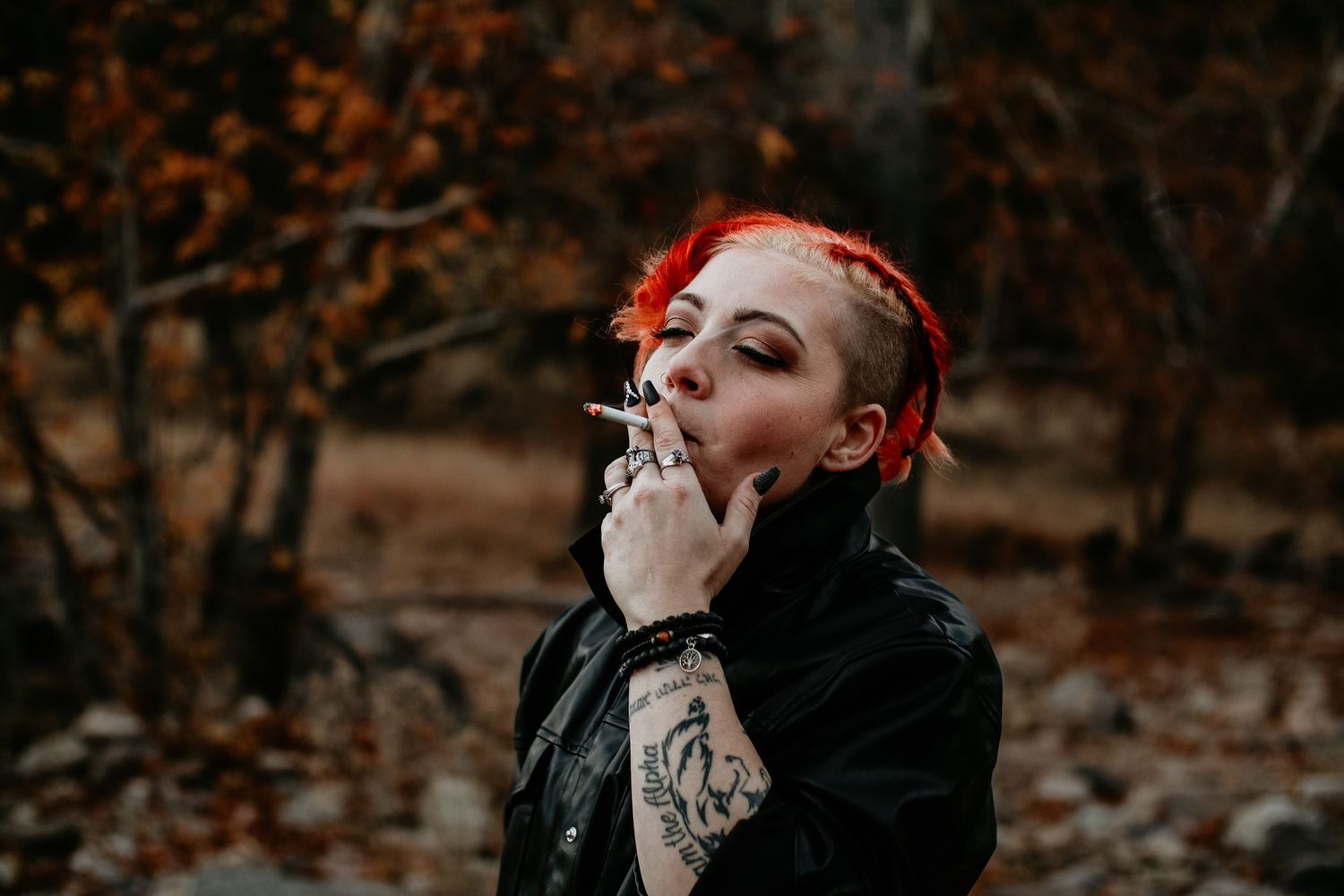 A young woman with red braided hair and tattoos wearing a black leather jacket smoking outdoors in autumn.