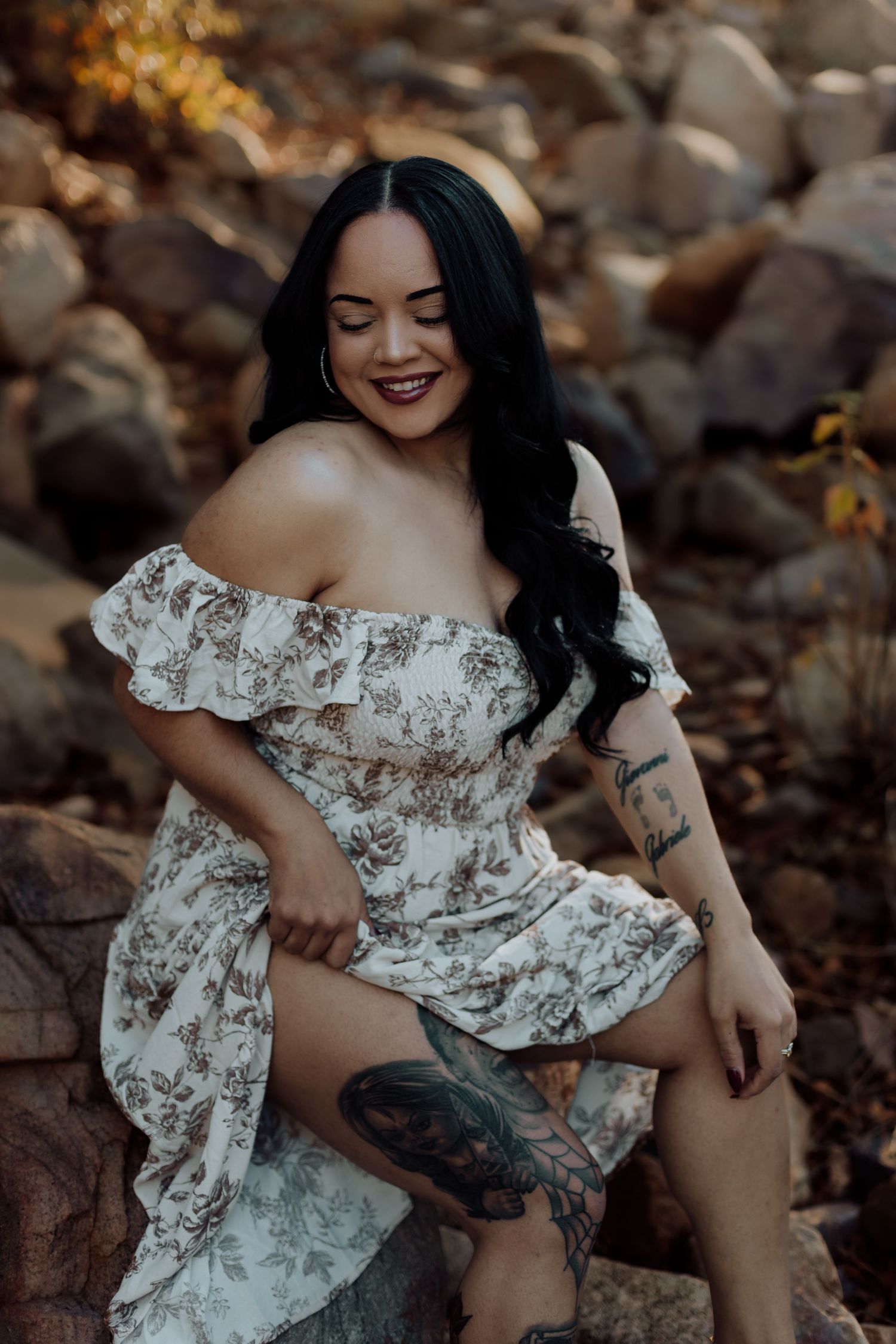 Smiling woman with black hair and tattoos wearing a floral off-shoulder dress, seated on rocks outdoors.