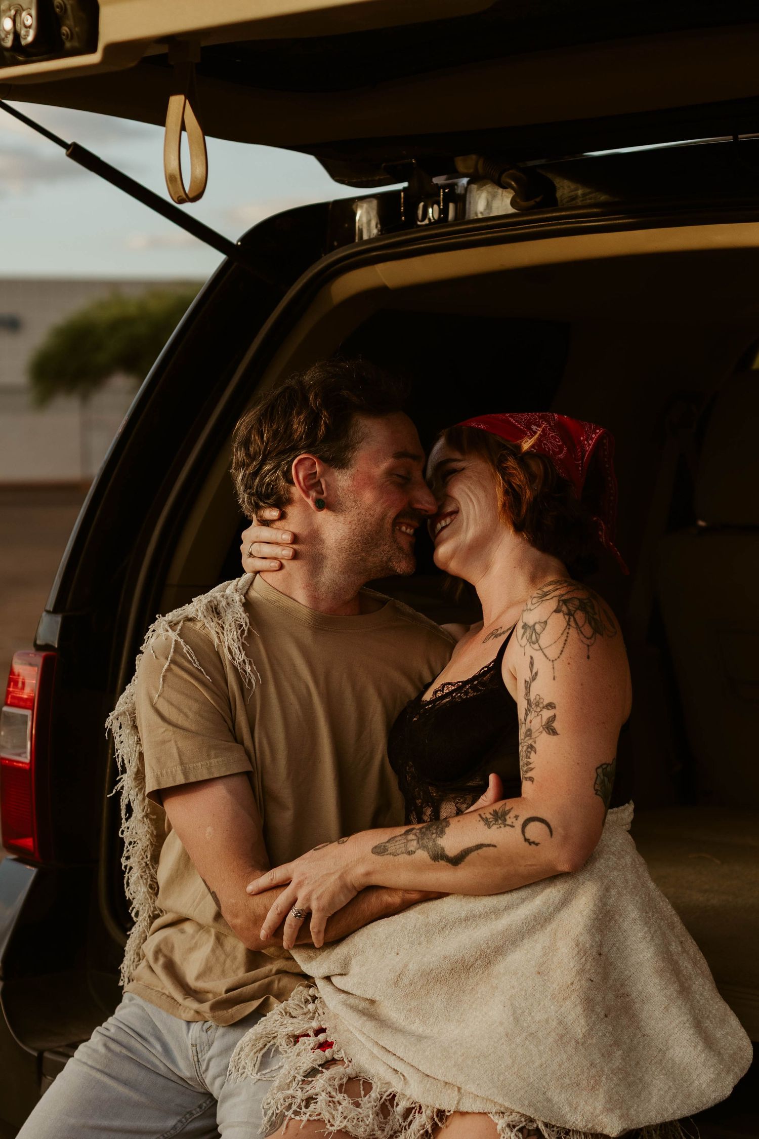 A couple sharing a joyful moment sitting in the trunk of an SUV, wrapped in a blanket at sunset.