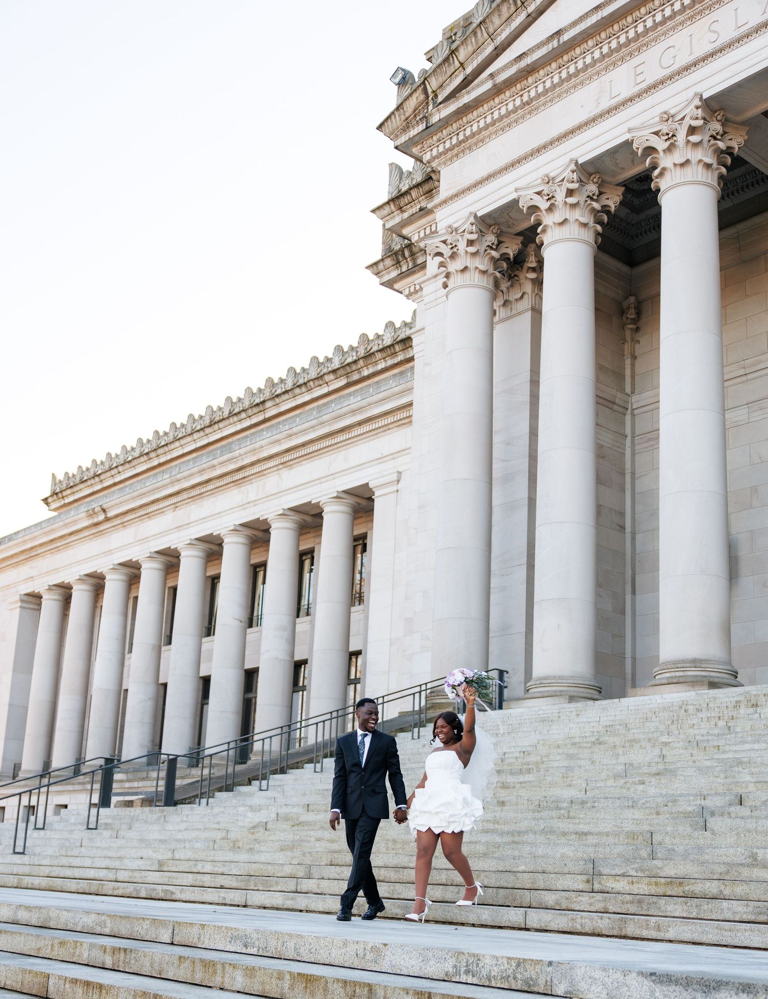 Newlyweds walking hand in hand beneath the columns of the Washington State Capitol