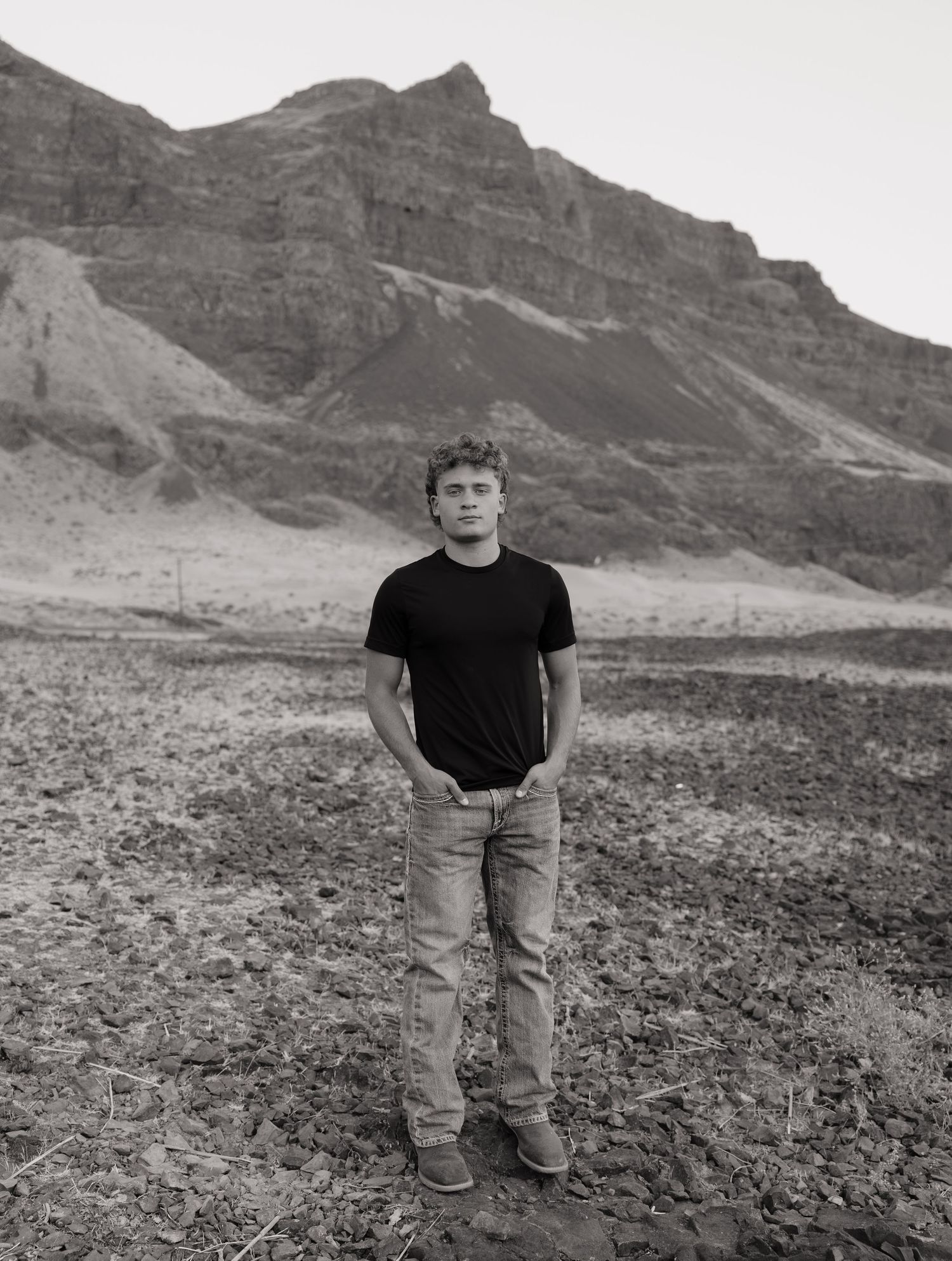 Black and white high school senior standing in a rocky desert landscape with dramatic cliffs behind him.