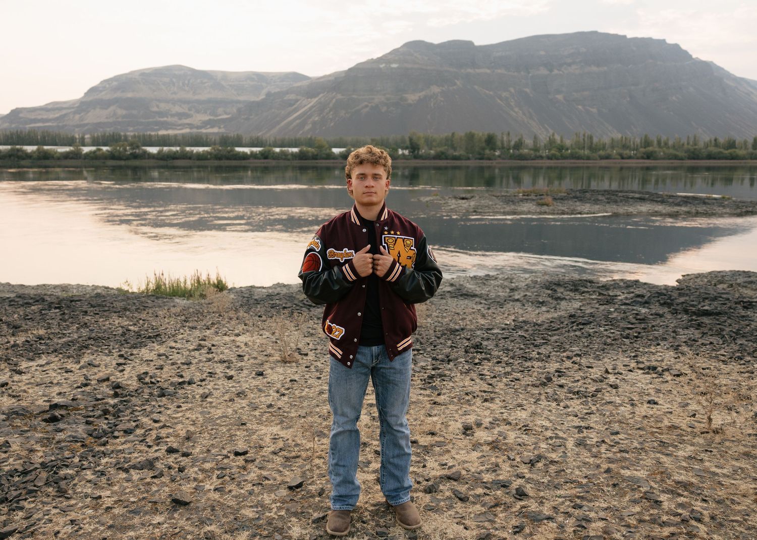 High school senior in a letterman jacket posing along the water with mountains in the background in Washington.