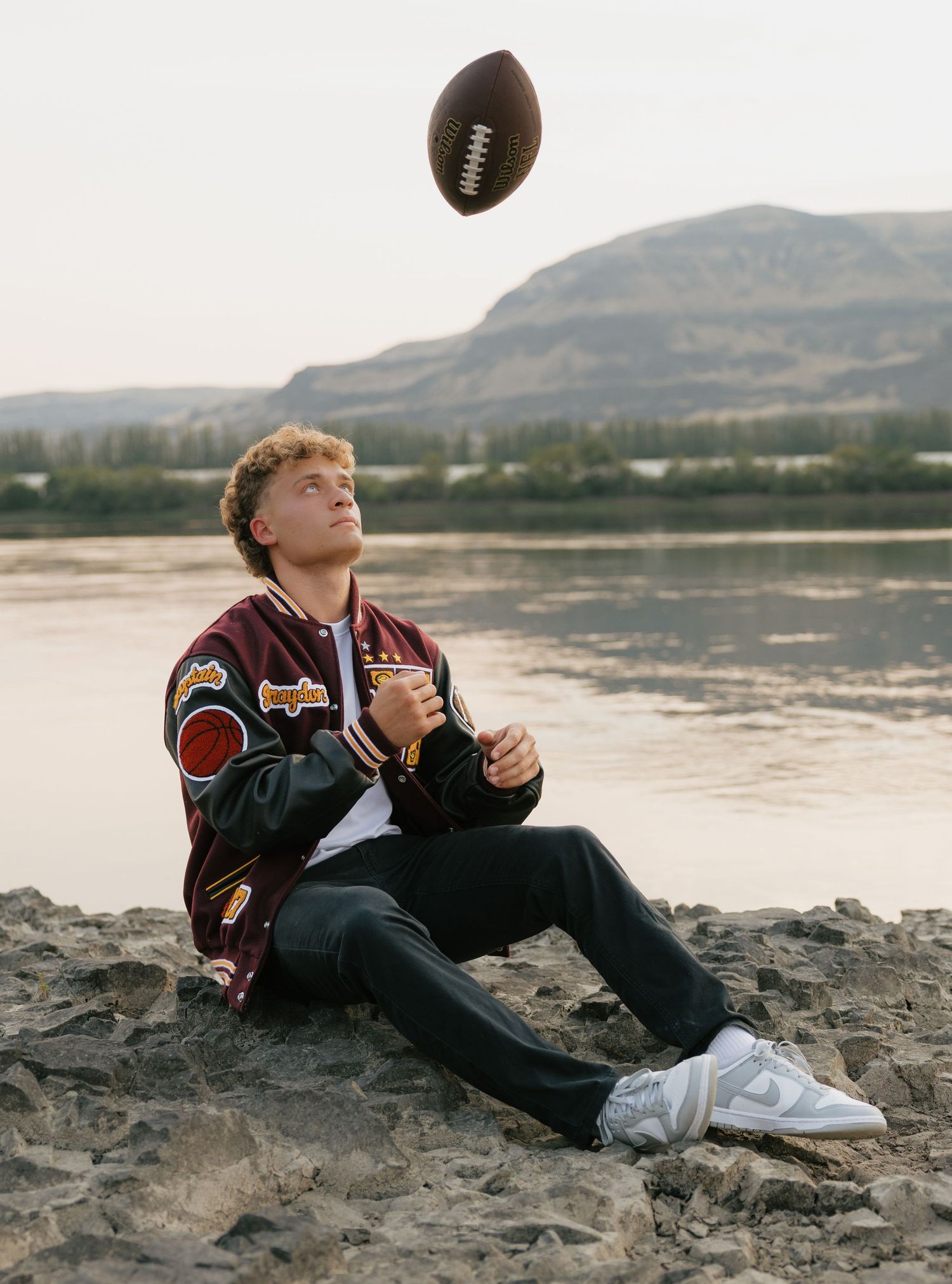 High school senior in a varsity jacket tossing a football during an outdoor senior photography session by the water.