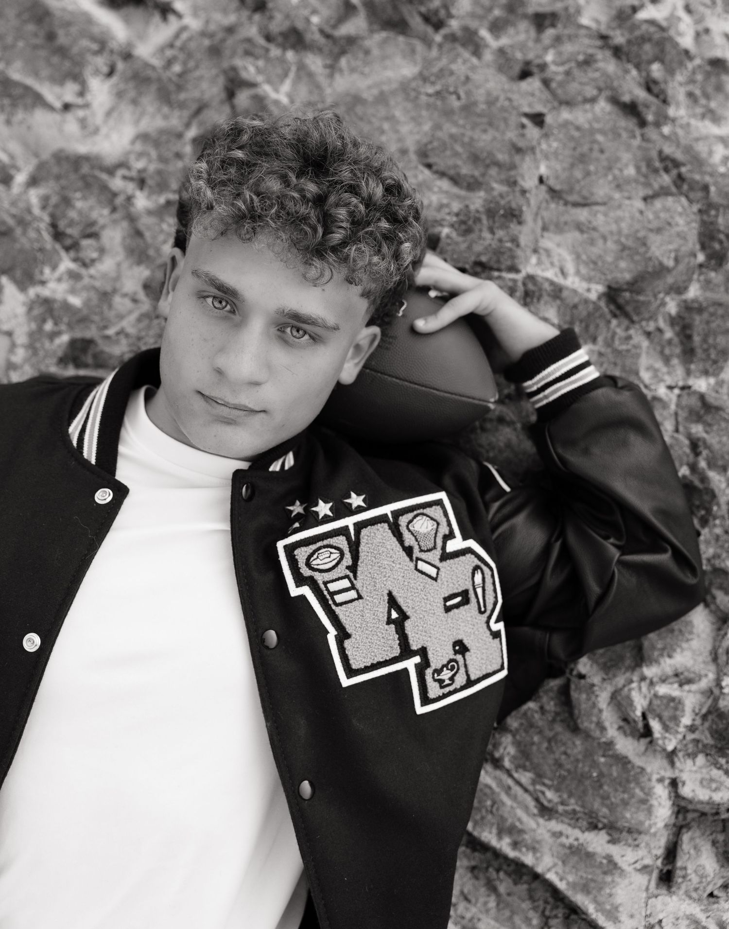 Black and white high school senior portrait in a varsity letterman jacket posing with a football against a textured rock wall.