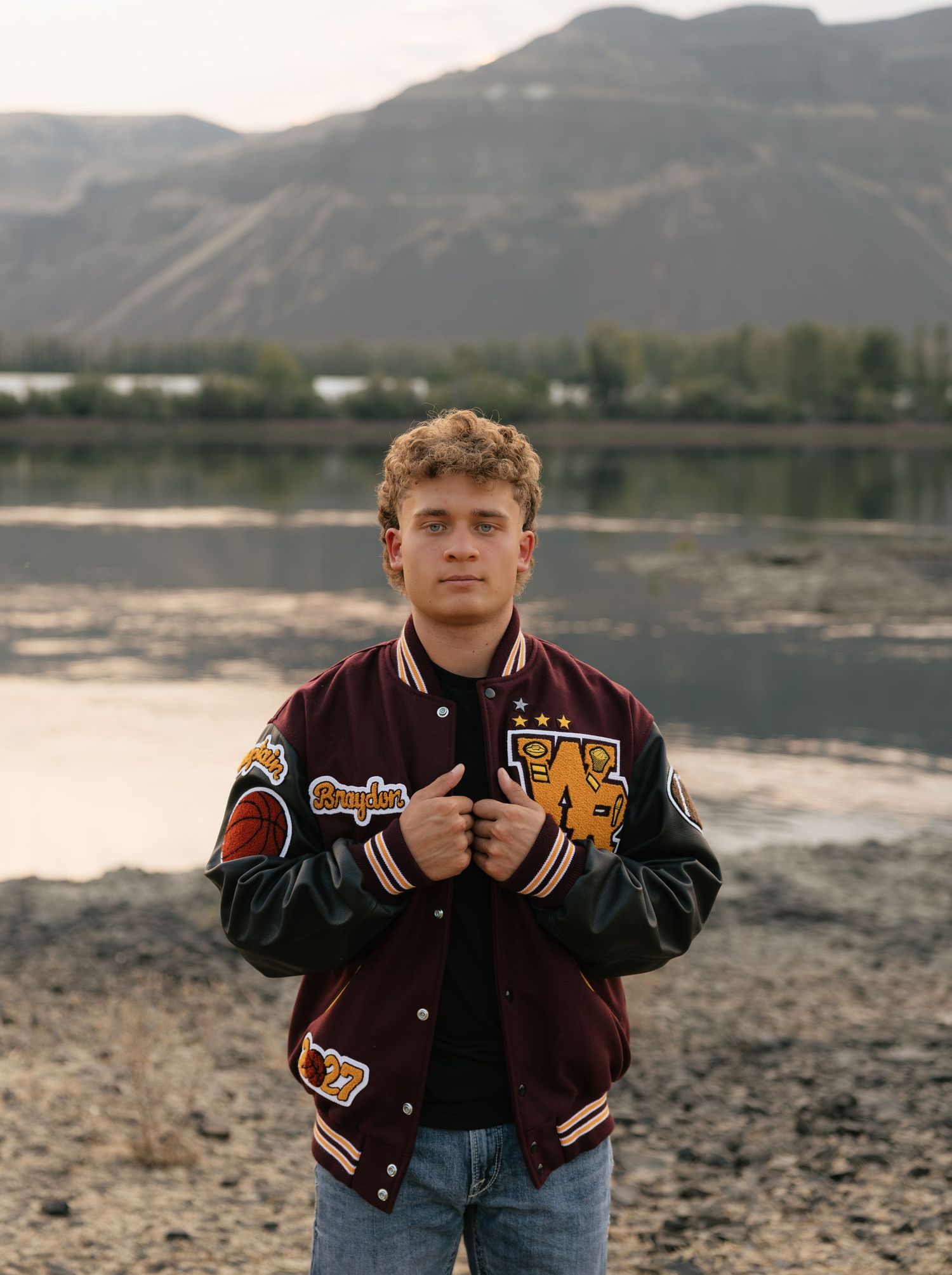 High school senior posing in a letterman jacket by the water with scenic Washington mountain views.
