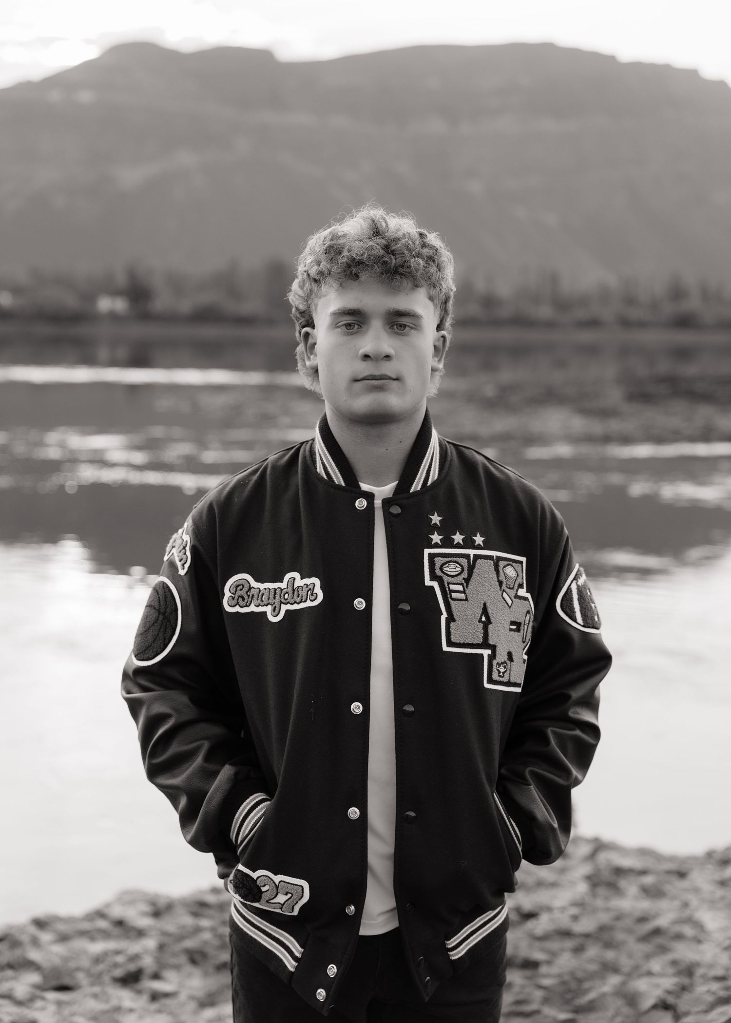 Black and white senior portrait in a varsity jacket by the water with scenic mountains in the background.