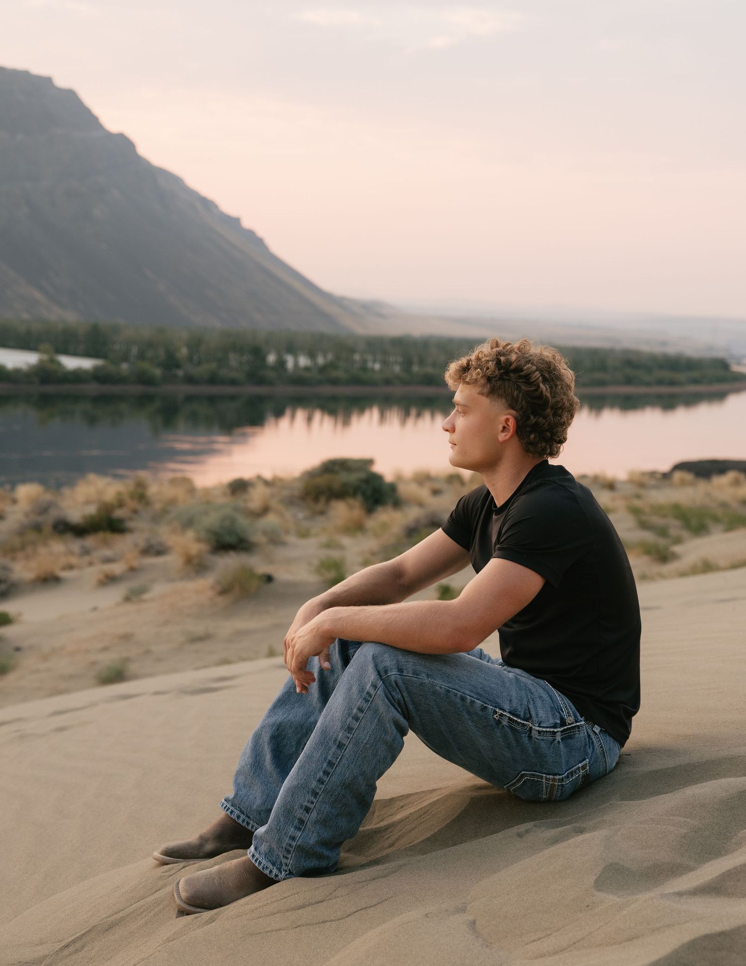 High school senior sitting on a sandy hillside overlooking the water and mountains during a golden hour senior session.
