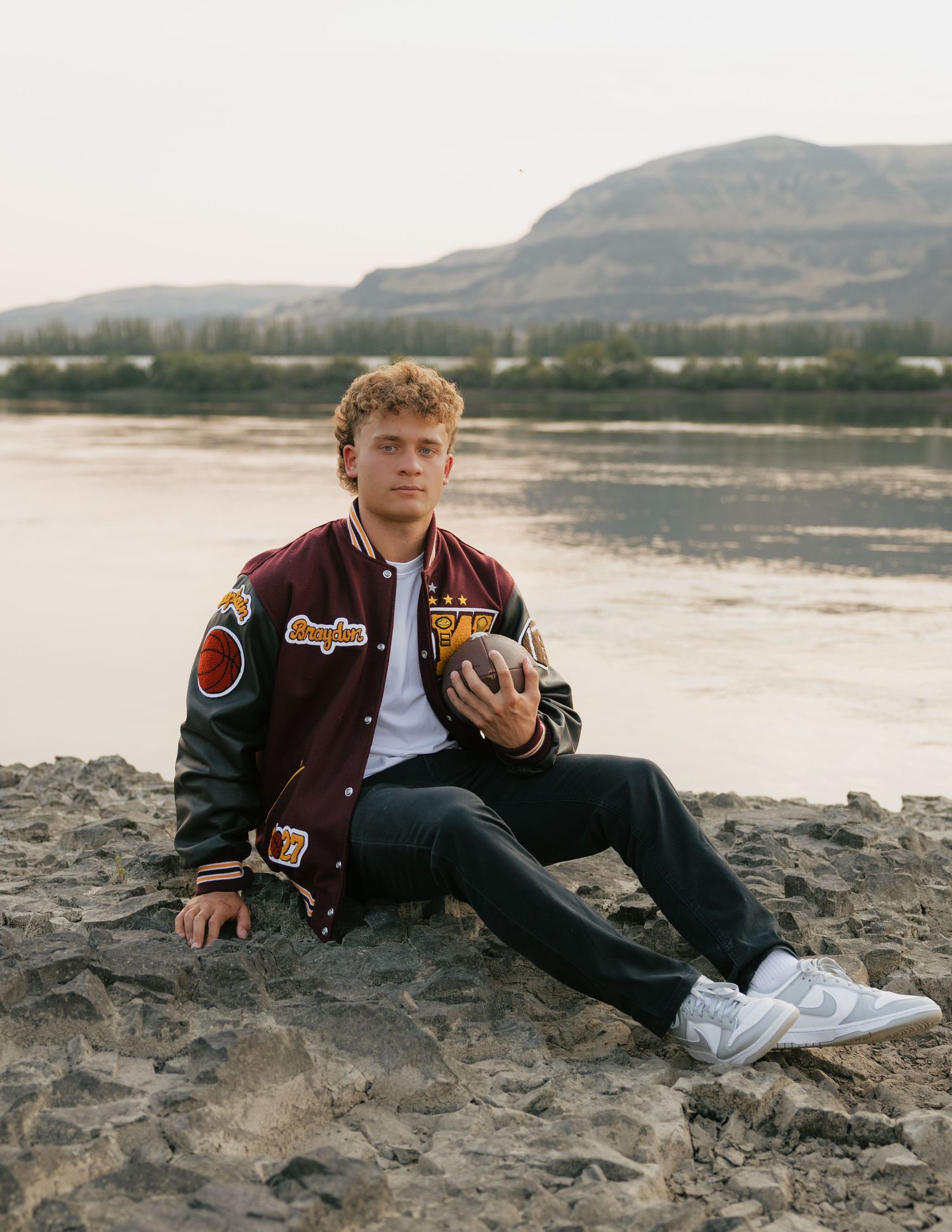 High school senior sitting along the river in his varsity letterman jacket holding a football with mountains behind him.