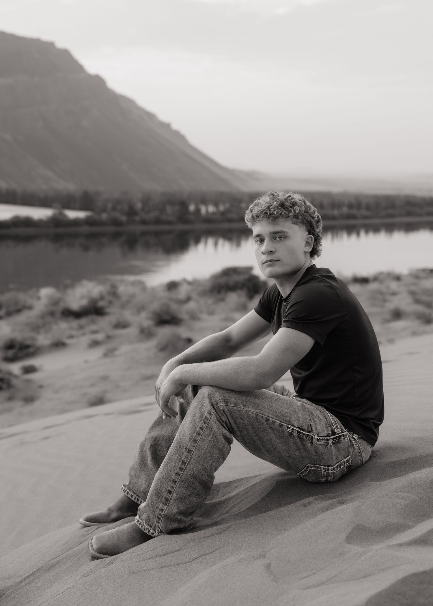 Black and white high school senior portrait sitting on sand beside the water with mountains in the background.