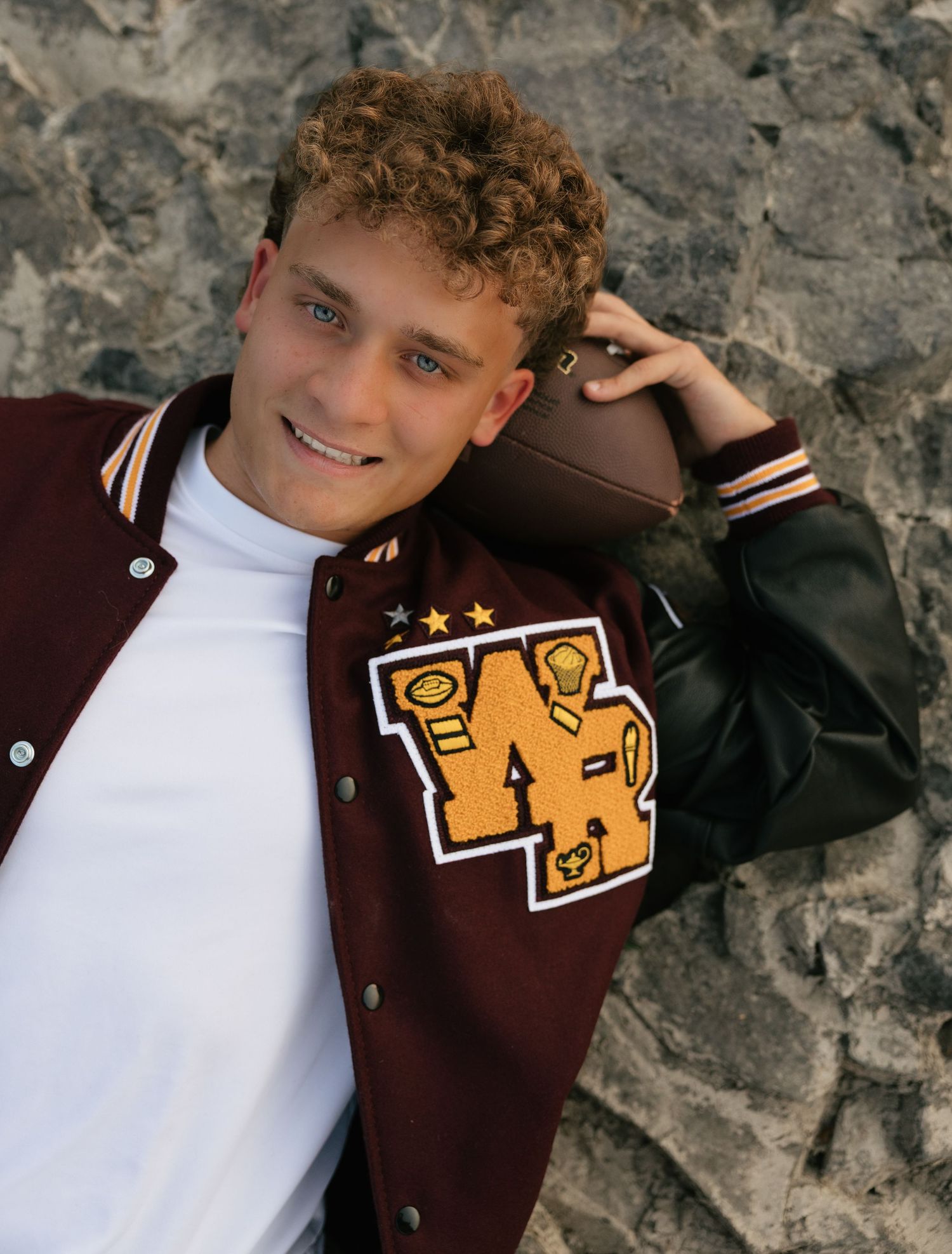 High school senior portrait in a varsity letterman jacket posing with a football during his outdoor senior session.