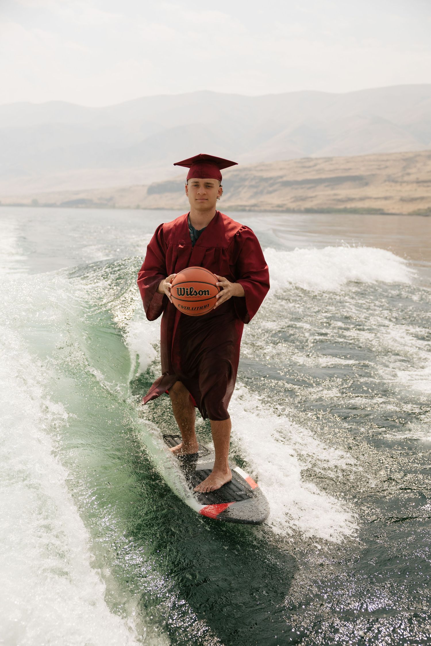 High school senior holding a basketball while wakesurfing in his graduation cap and gown on the lake.
