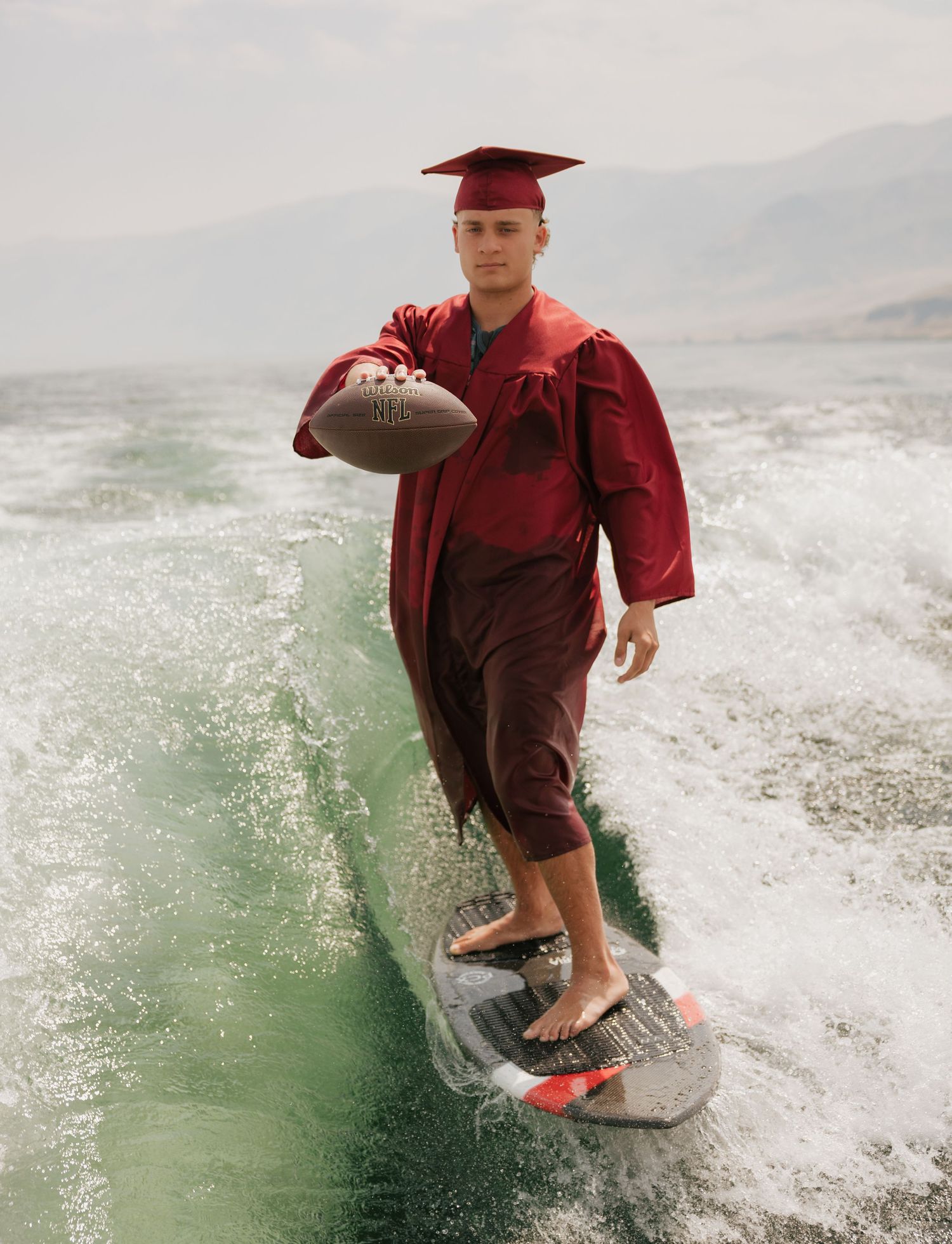 High school graduate holding a football while wakesurfing in his cap and gown during a unique senior session.