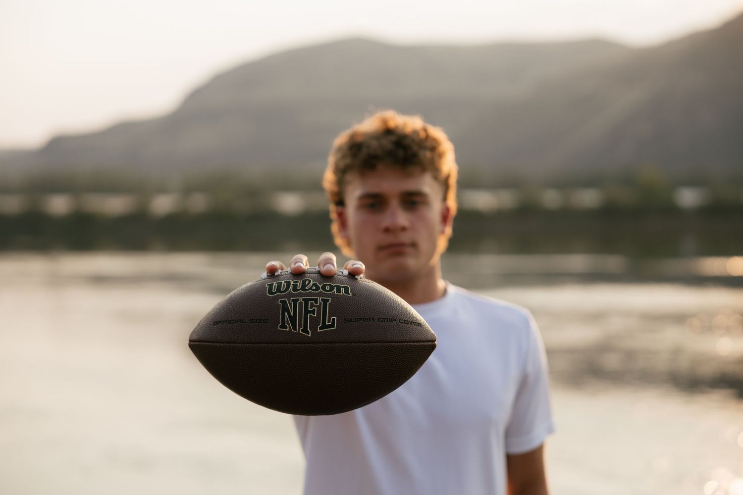 High school senior holding a football toward the camera with scenic water and mountains in the background.