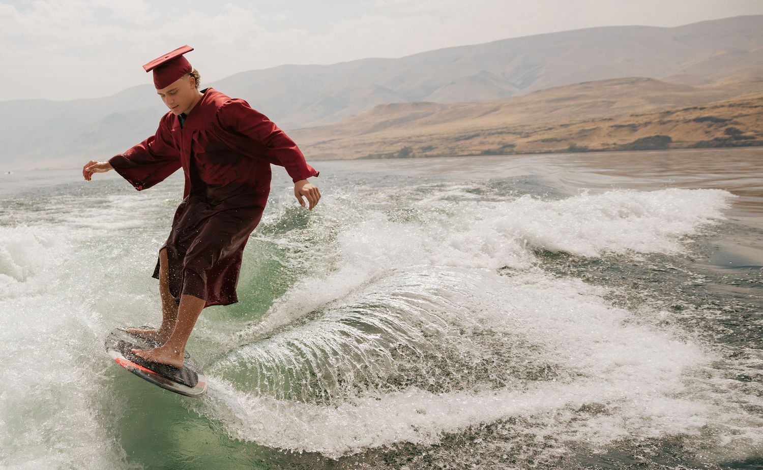 High school graduate wakesurfing in a cap and gown during a creative outdoor senior photography session.