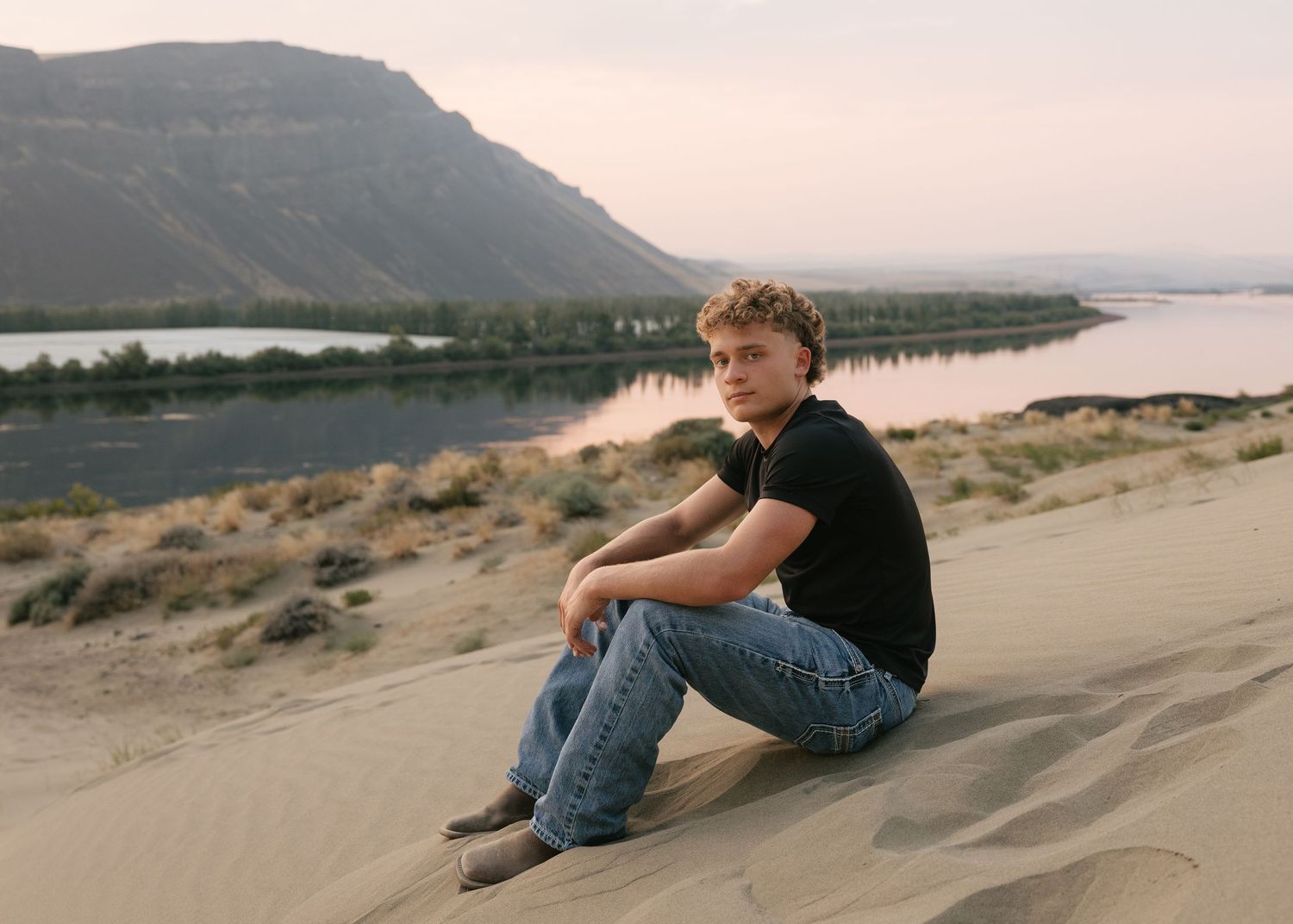 High school senior sitting on a sandy hillside overlooking the water and mountains during a golden hour senior session.