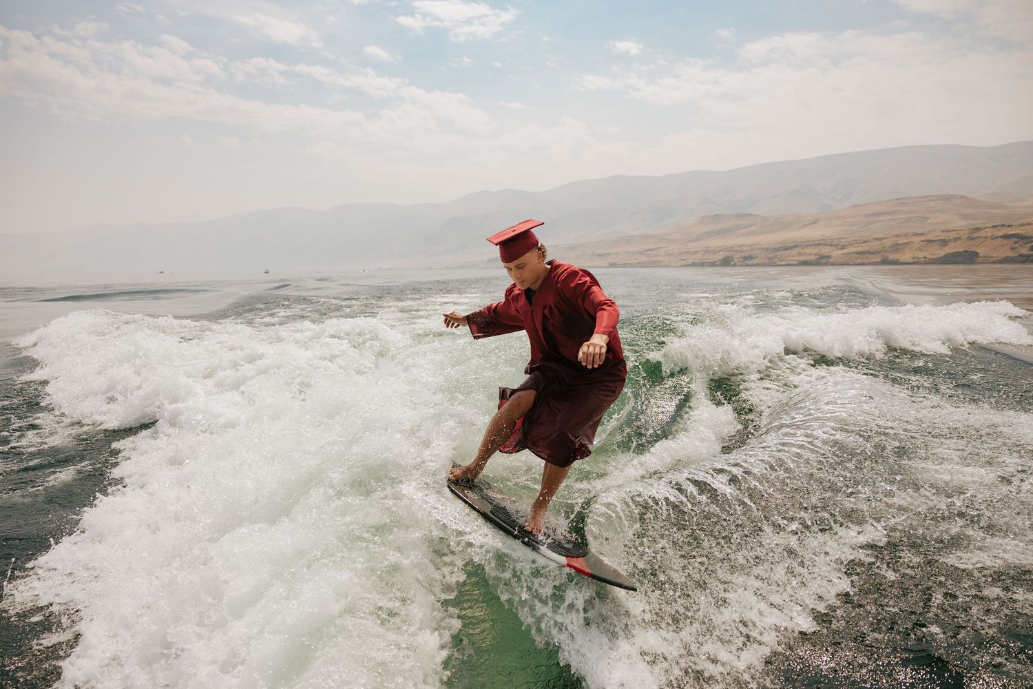 High school senior wakesurfing in his graduation cap and gown with scenic hills in the background.