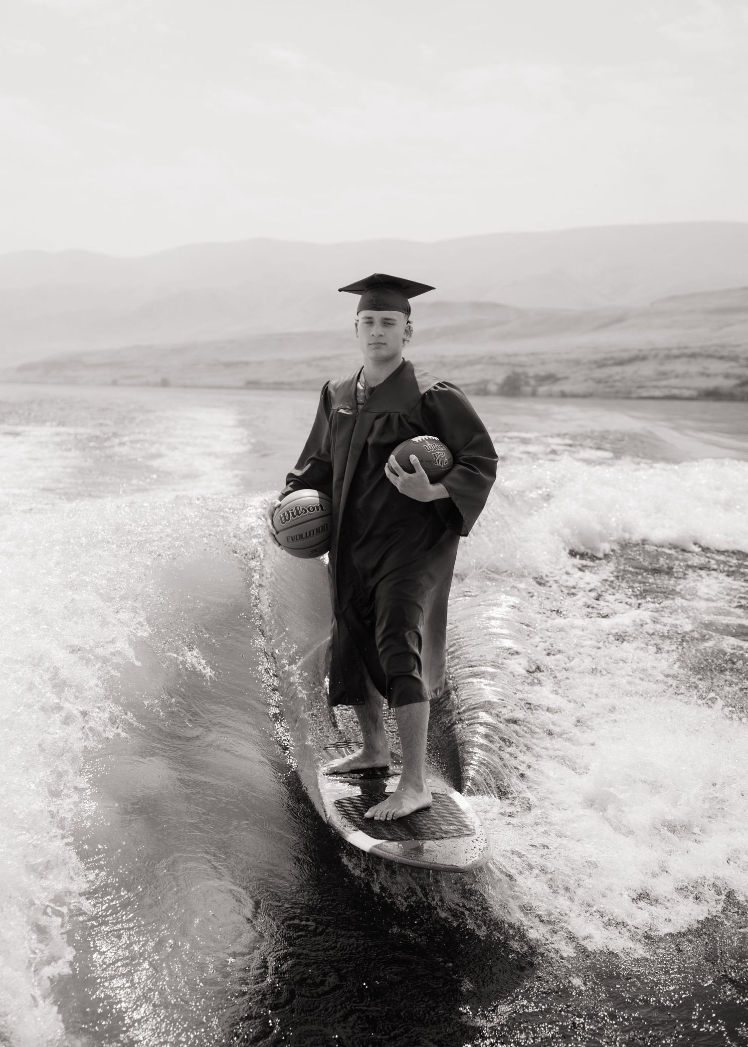 Black and white senior portrait of a graduate wakesurfing in his cap and gown on a Washington lake.