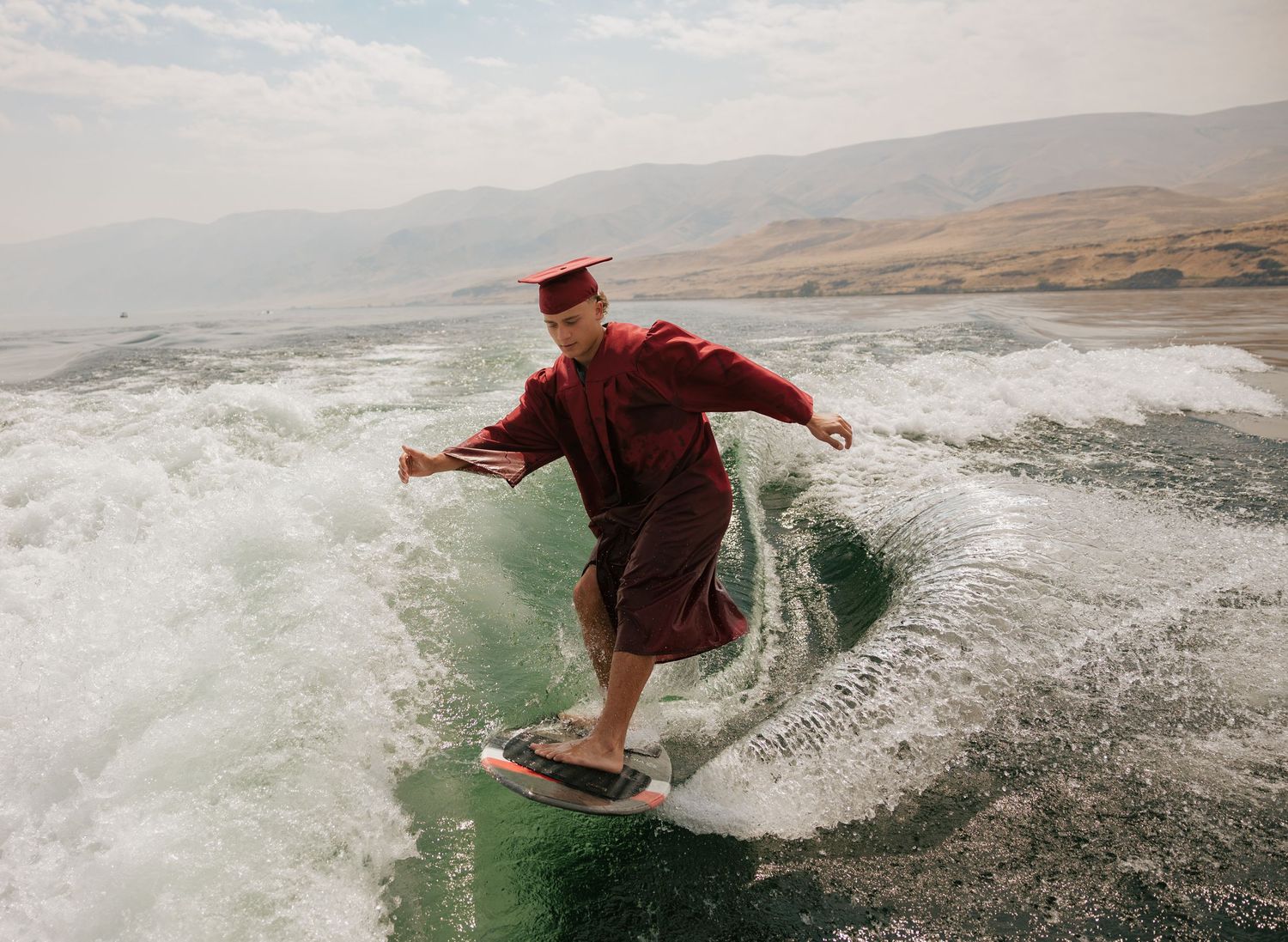 Creative senior portrait of a graduate wakesurfing in his cap and gown on the lake.