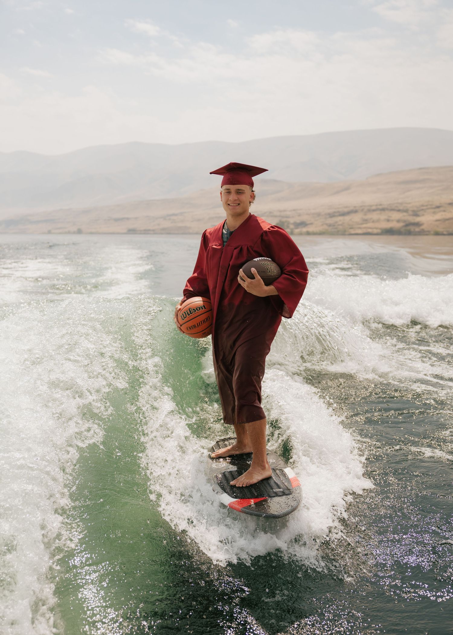 High school graduate wakesurfing in a cap and gown while holding football and basketballs during a creative senior session.