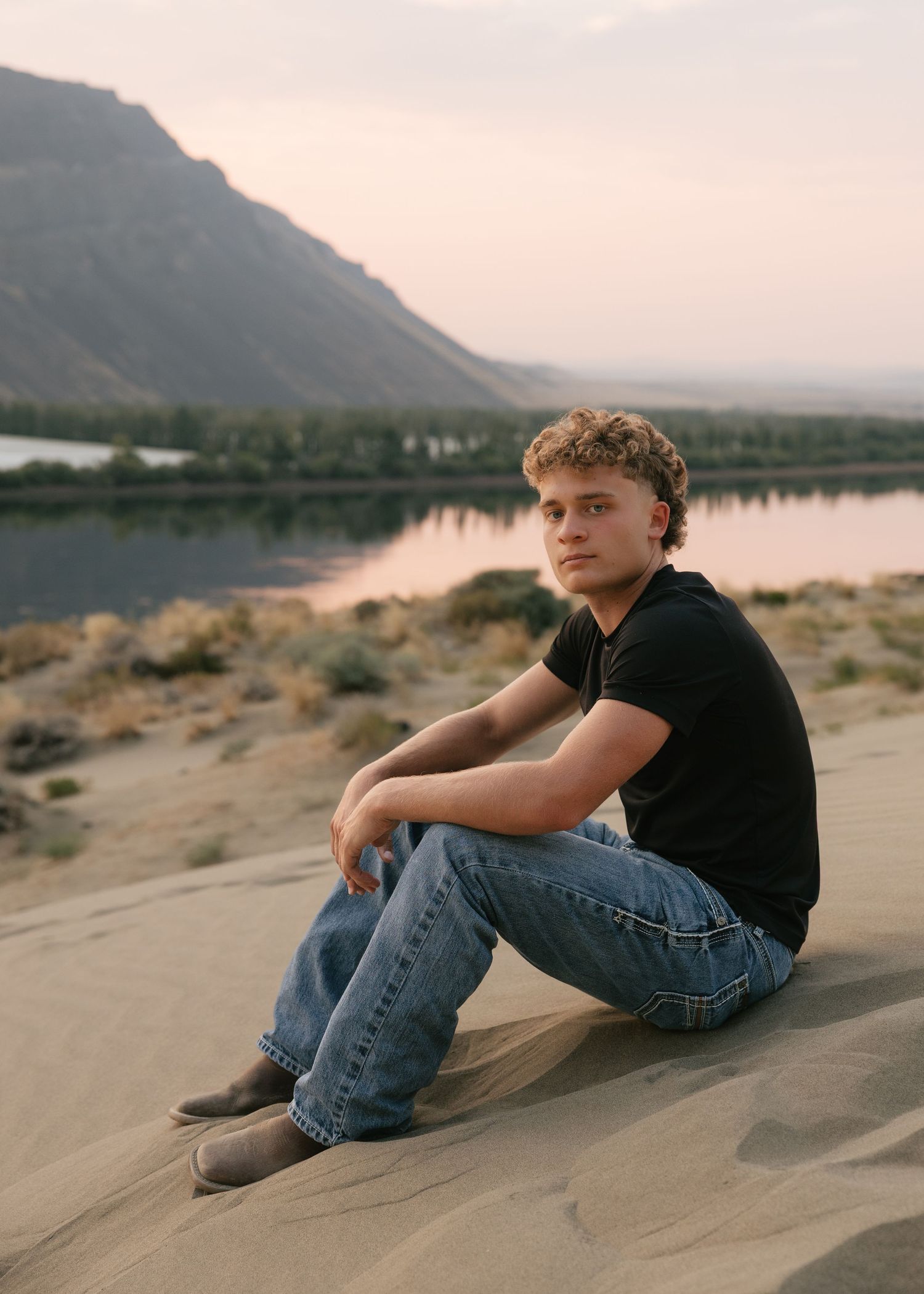 High school senior sitting on the sand beside the water with mountain views during a golden hour senior session.