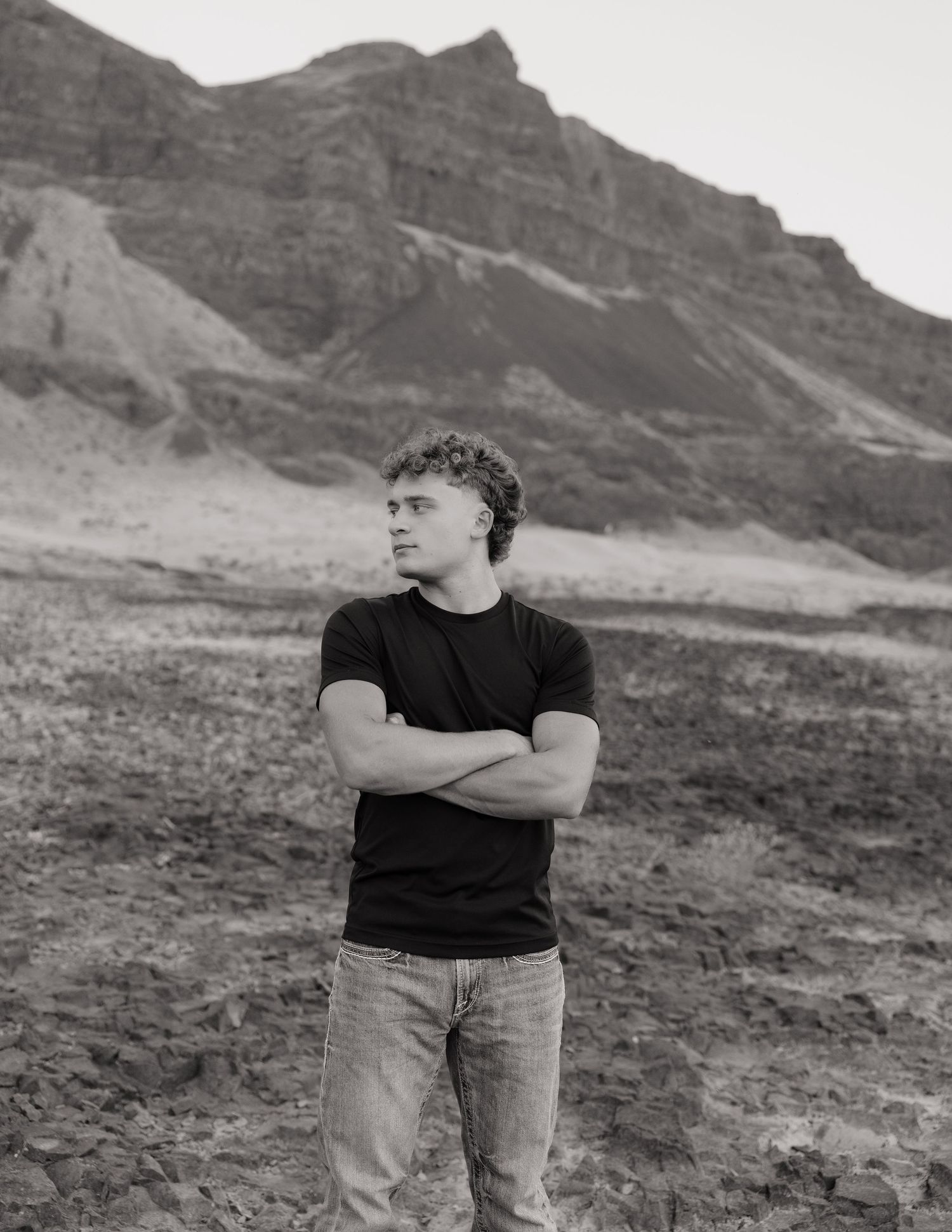 Black and white senior portrait standing with arms crossed in a rugged mountain landscape.