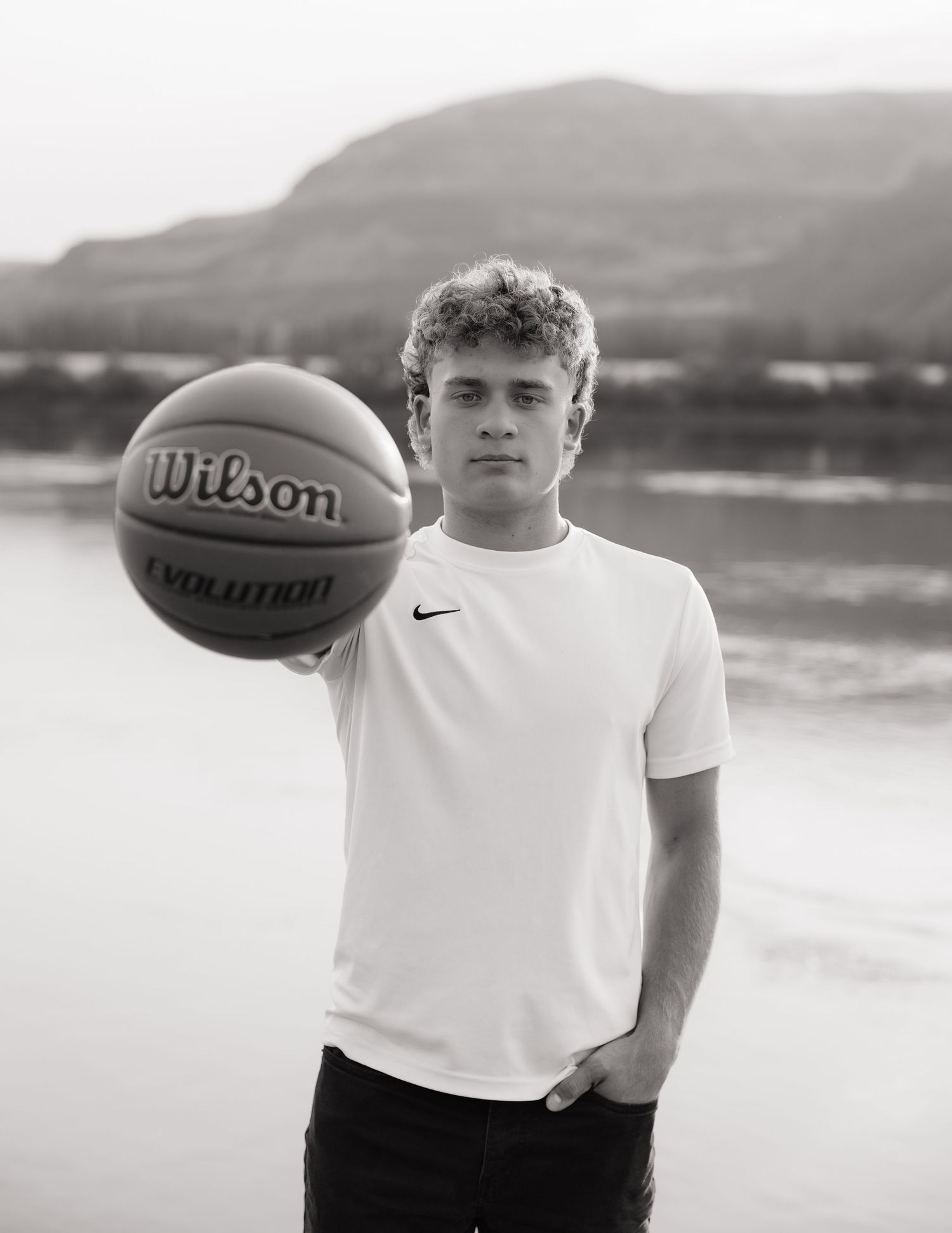 Black and white high school senior basketball portrait by the water with mountains in the background.