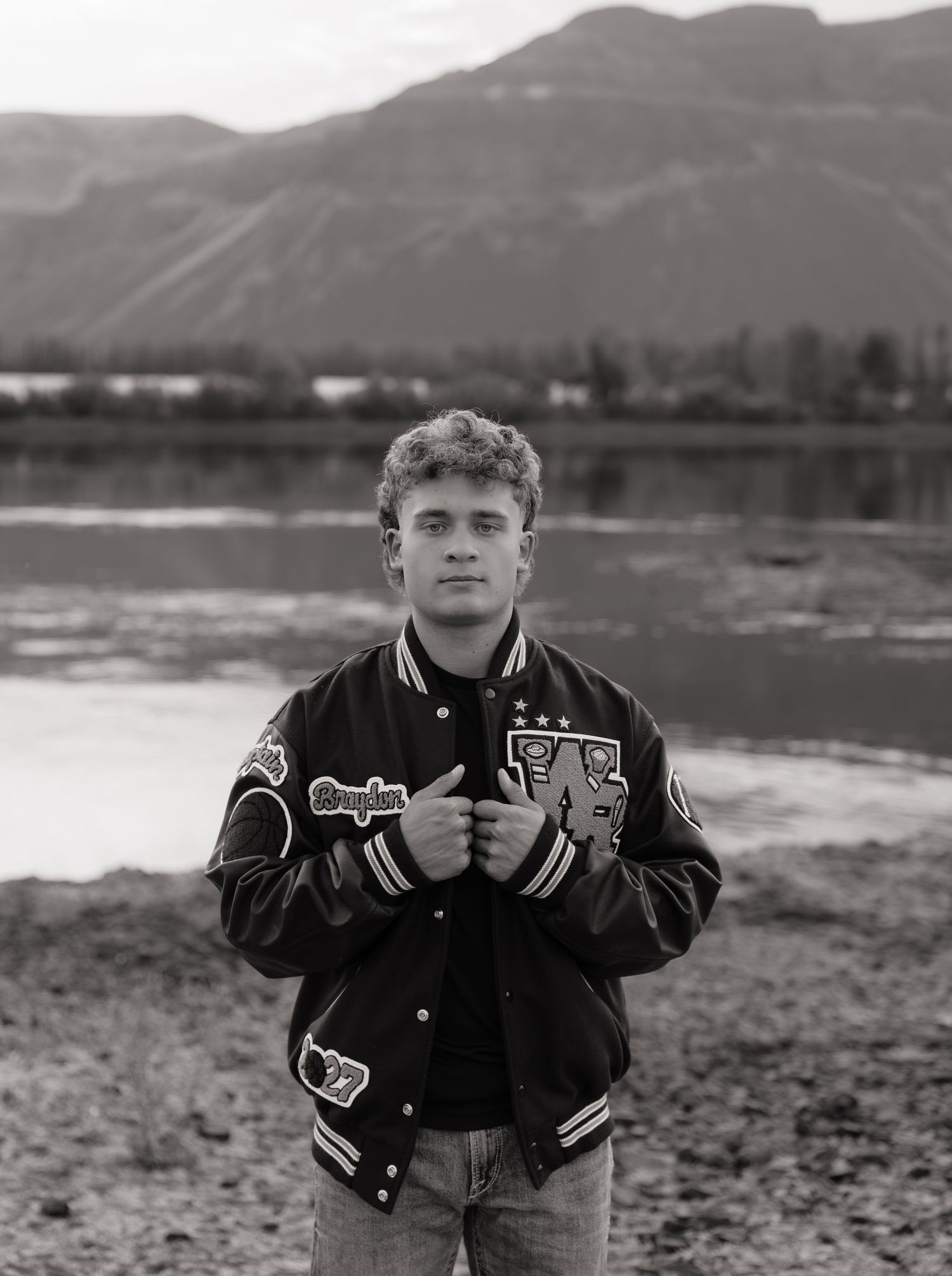 Black and white senior portrait in a letterman jacket with scenic mountains and water in the background.