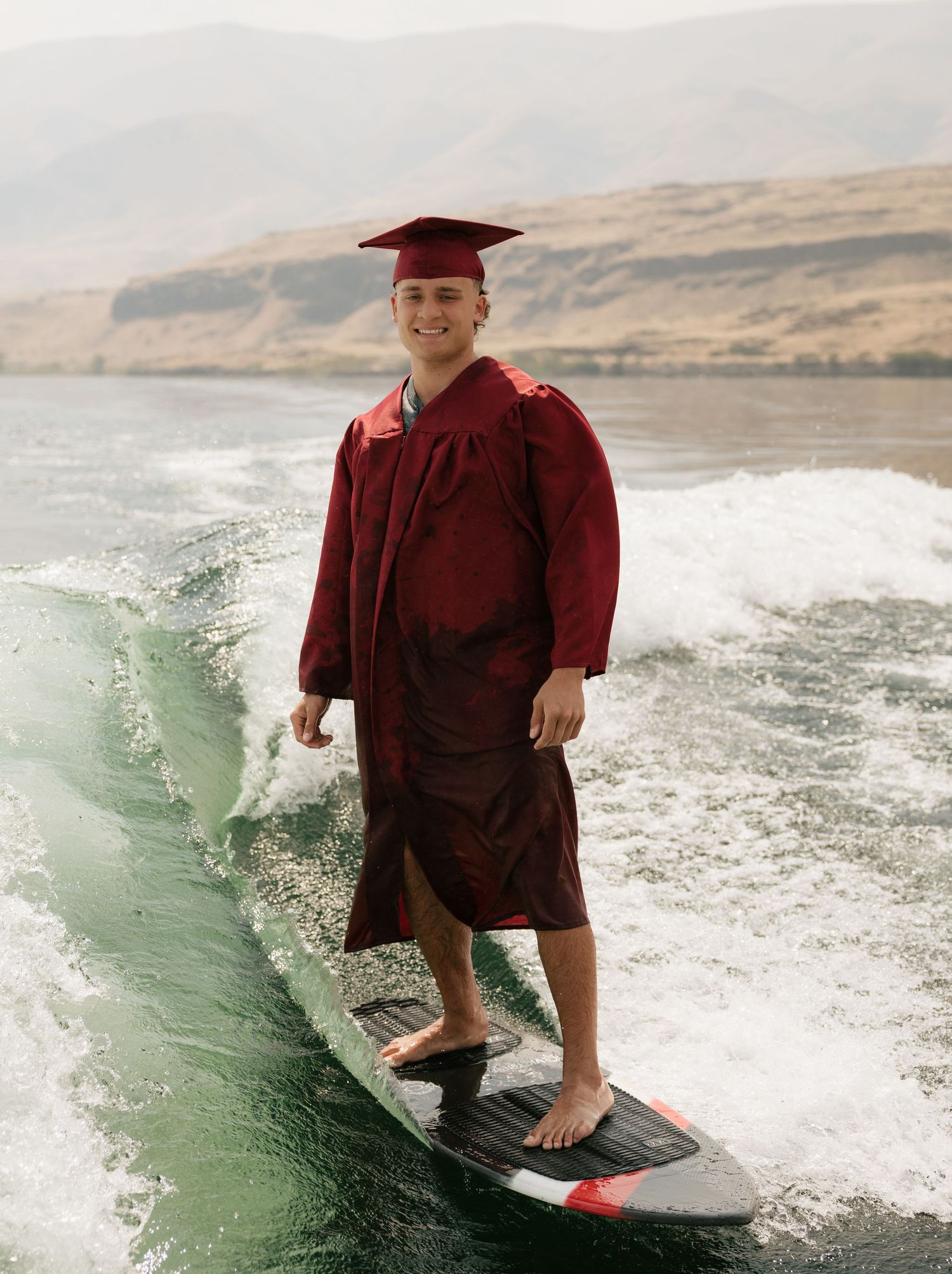High school graduate smiling while wakesurfing in his cap and gown during a unique outdoor senior session.