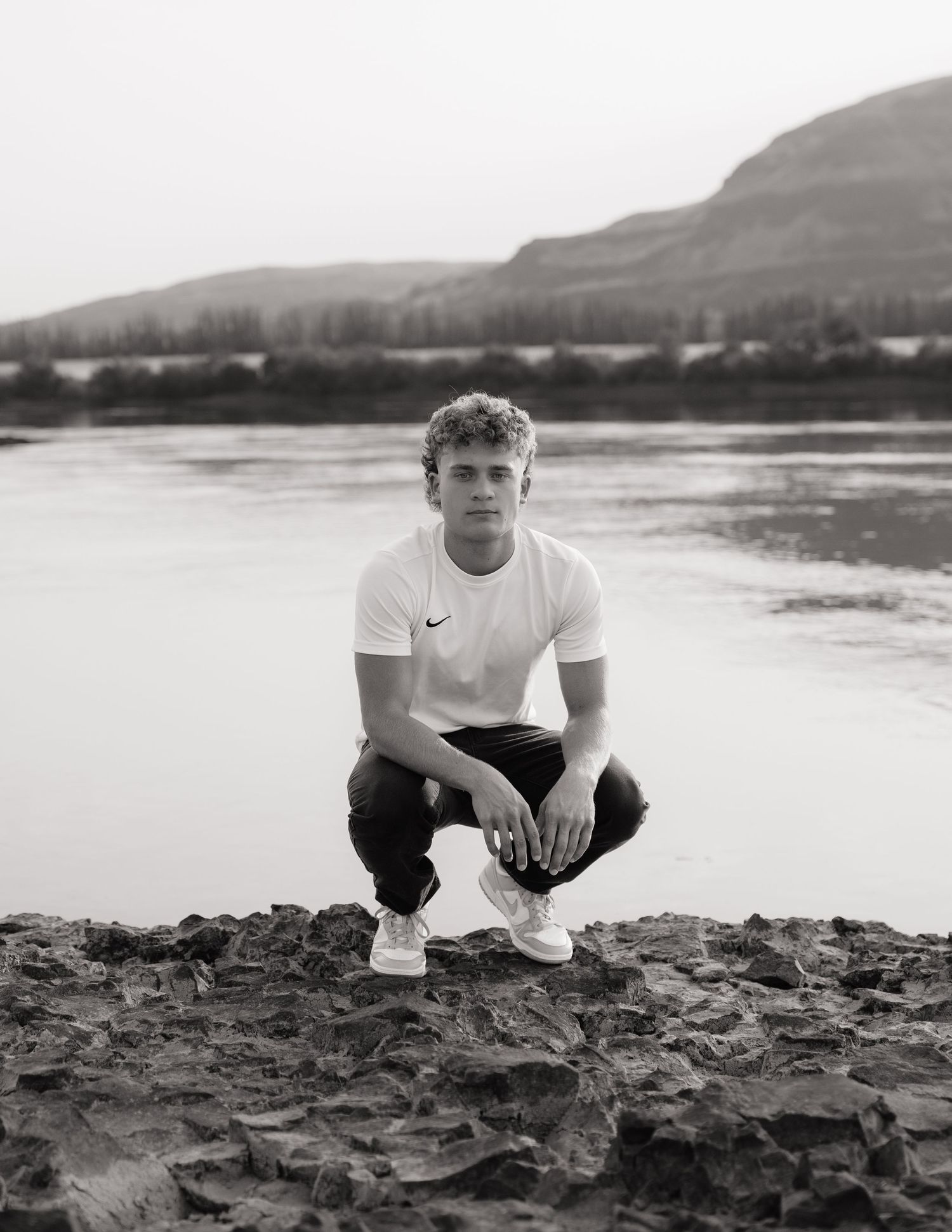 Black and white senior portrait crouching along the shoreline with water and mountain views behind him.