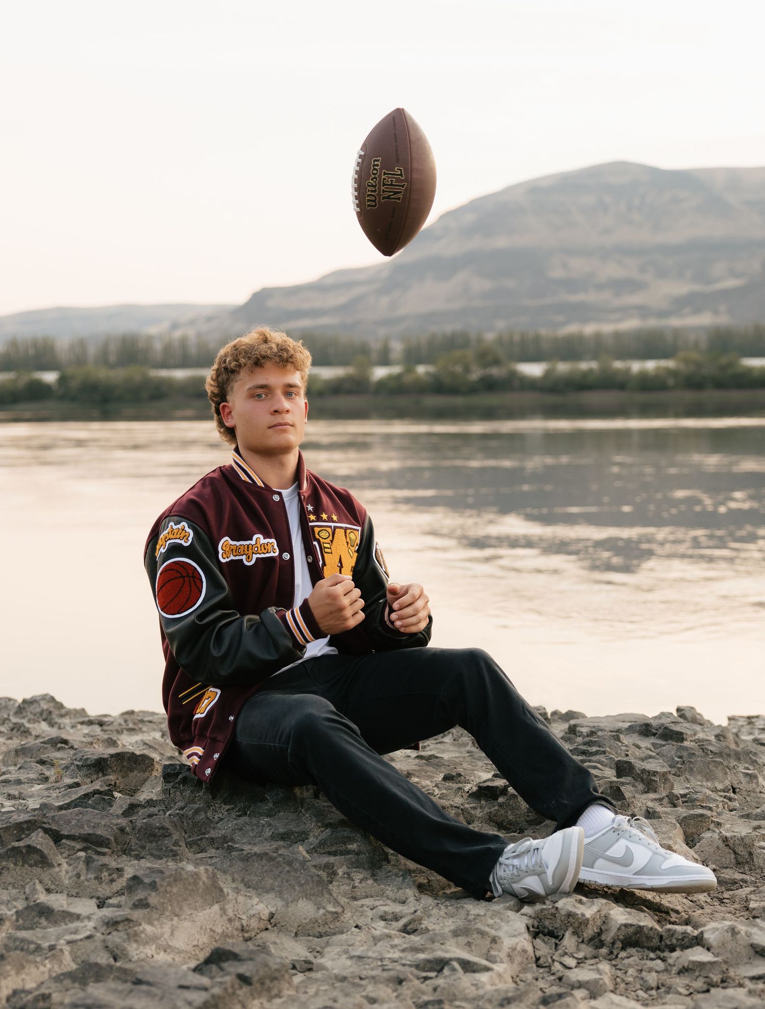 High school senior tossing a football while wearing his varsity letterman jacket beside the river and mountains.