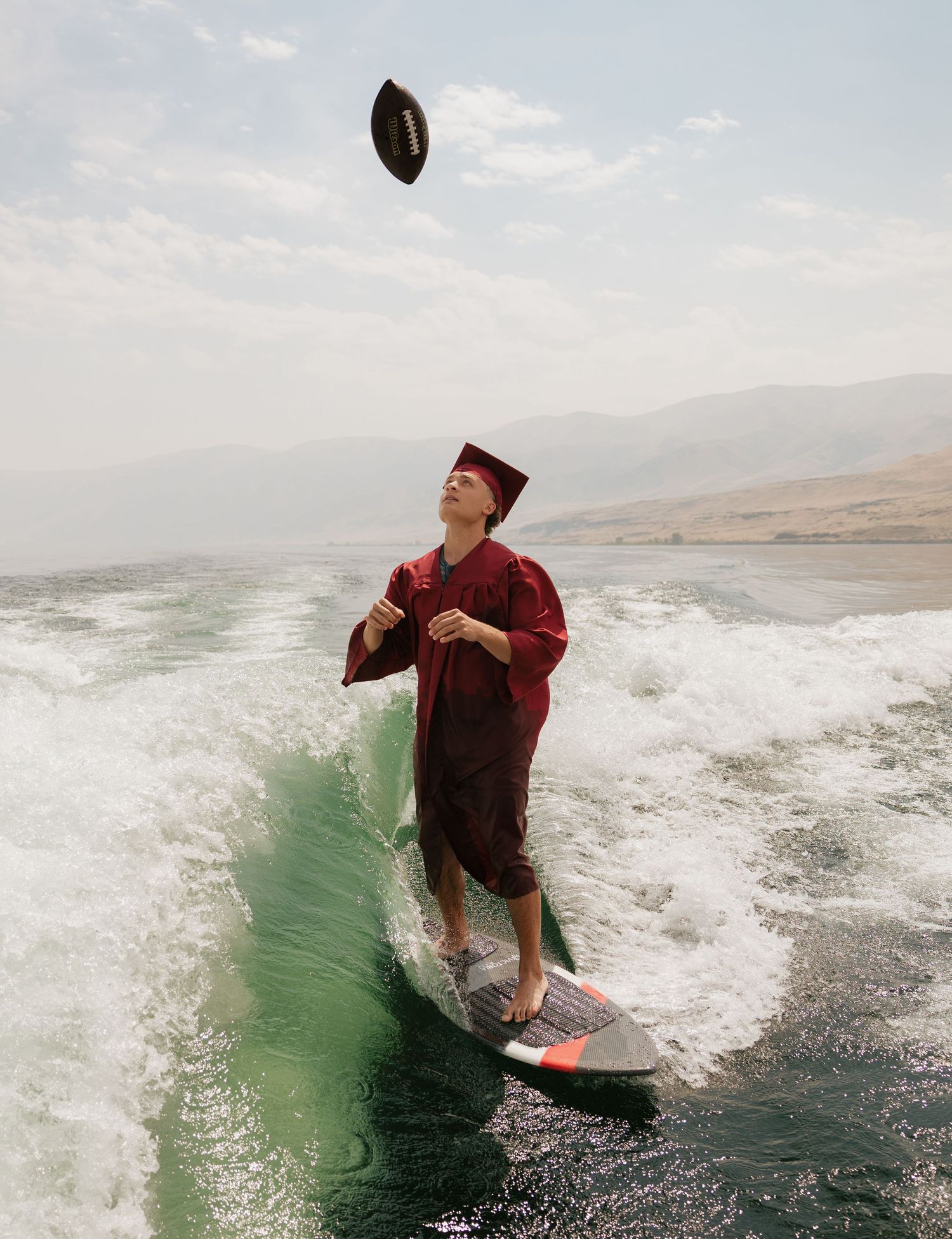 High school graduate tossing a football while wakesurfing in his cap and gown during a creative senior session.