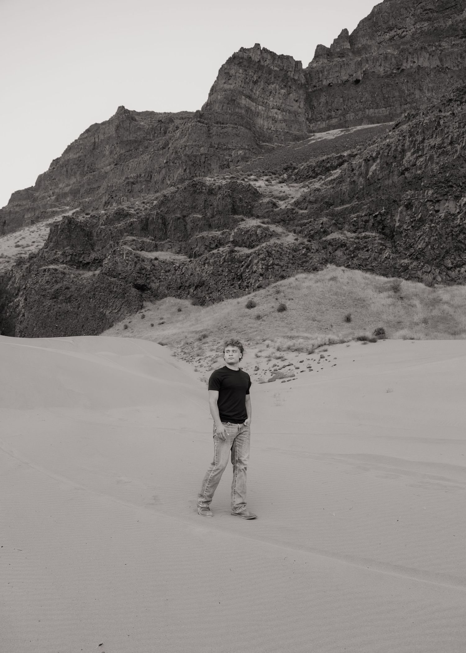 Black and white senior portrait in a dramatic desert landscape surrounded by rugged mountain cliffs.