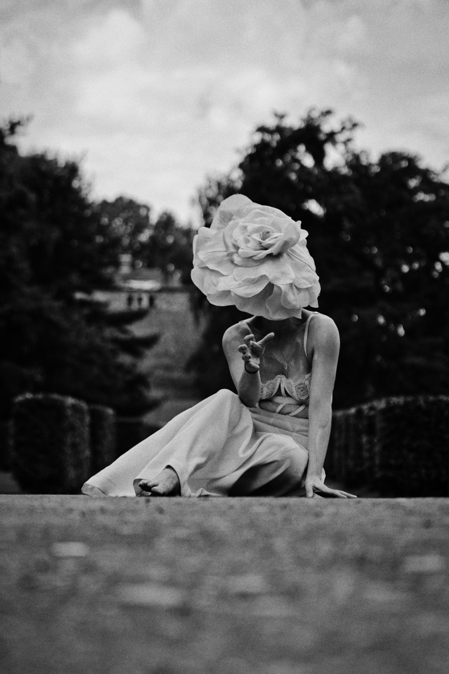 Black and white photo of a woman seated on a garden path, her face hidden by an oversized rose-shaped hat, with trees behind.