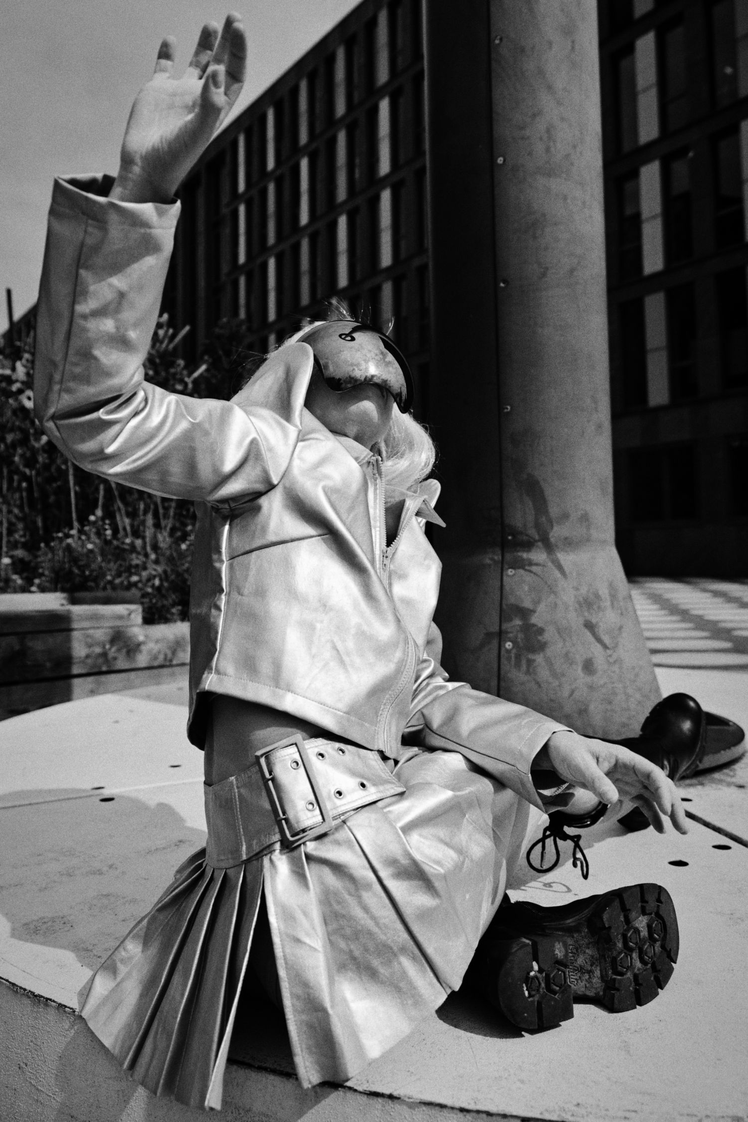 Black and white photo of a person in a metallic trench coat, pleated skirt and mask kneeling by a stone pillar, one arm raised.