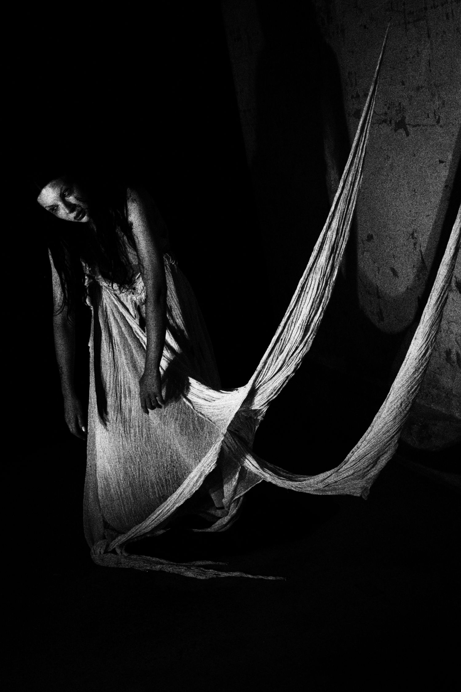 Black and white photo of a woman draped in sheer fabric emerging from darkness in an ethereal, haunting pose.