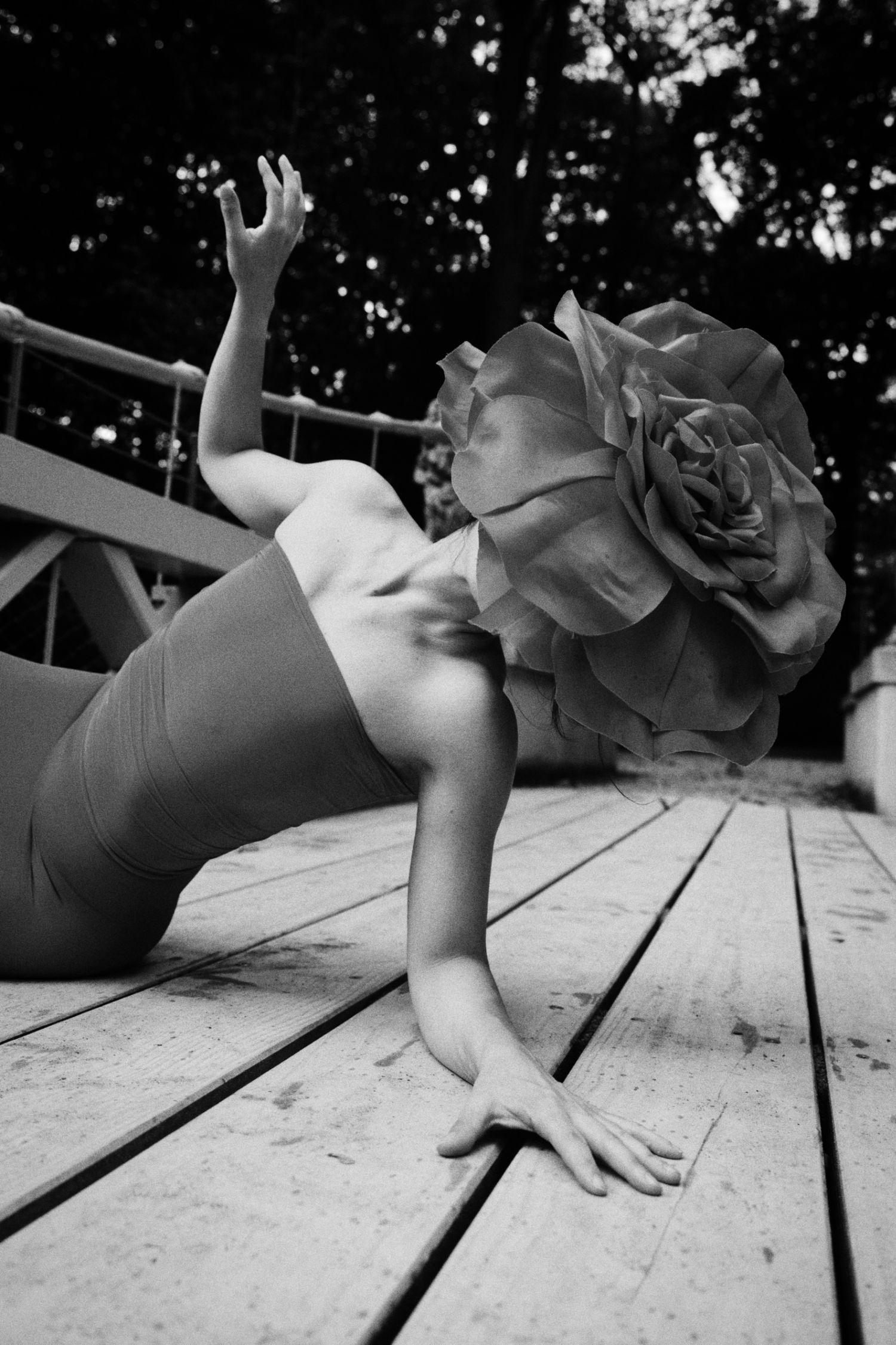Black and white photo of a woman in a strapless dress reclining on a wooden deck, her head hidden by a giant paper rose, one hand raised.