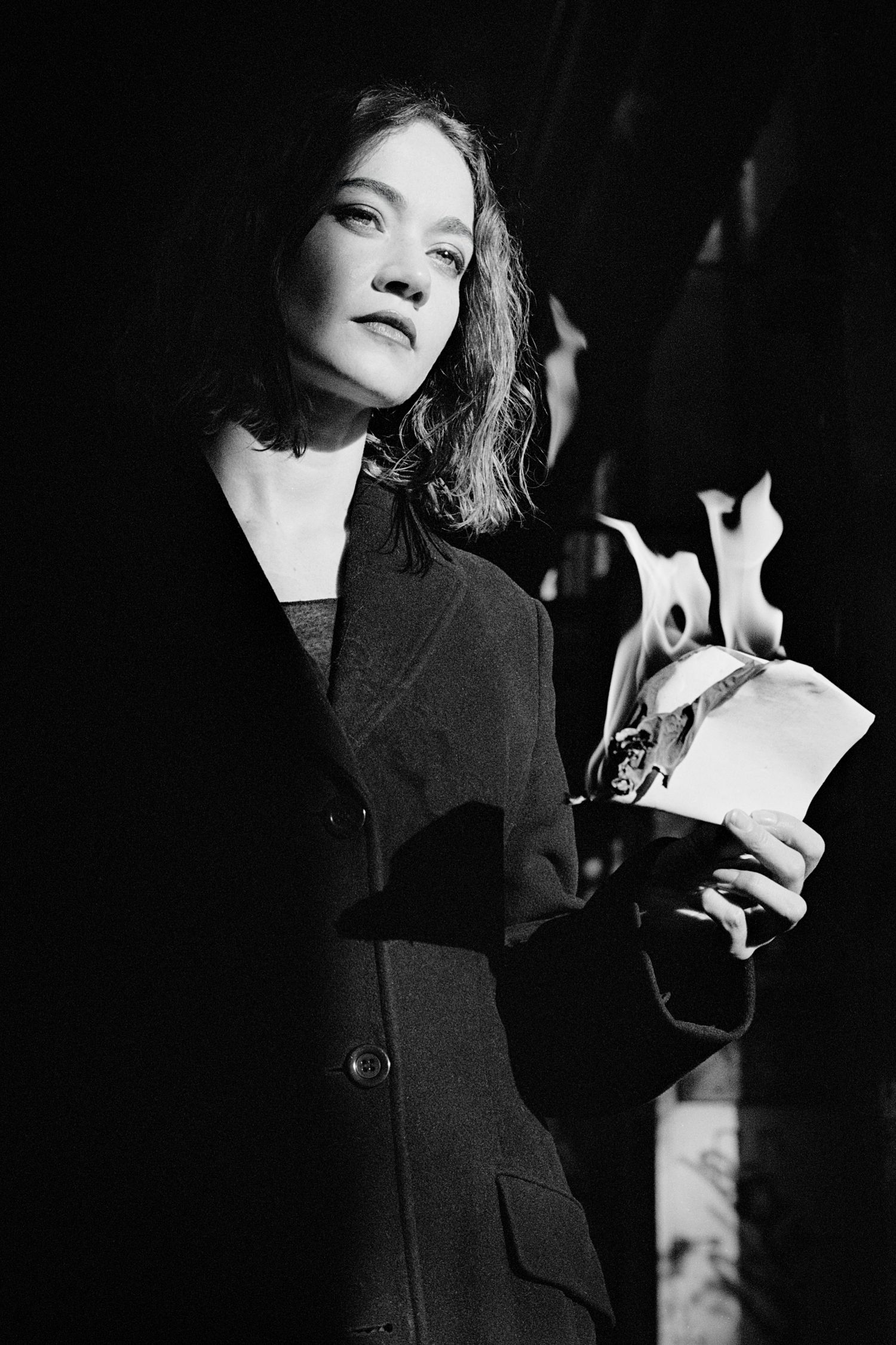 Black and white photo of a woman in a dark coat holding a burning book, dramatic lighting.