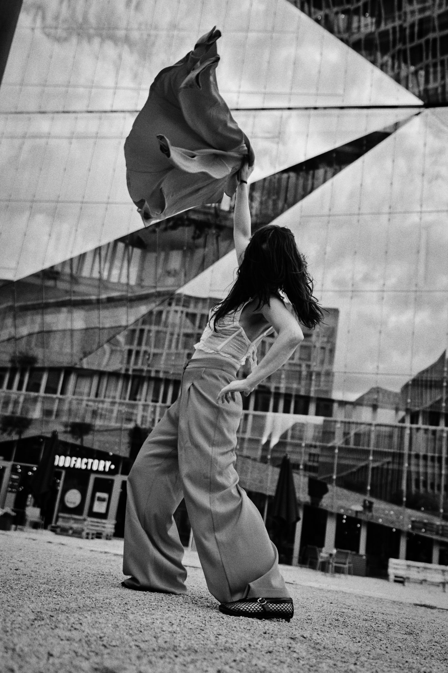 Black and white photo of a dancer in wide-leg trousers swirling a flowing fabric in front of a mirrored geometric glass building.