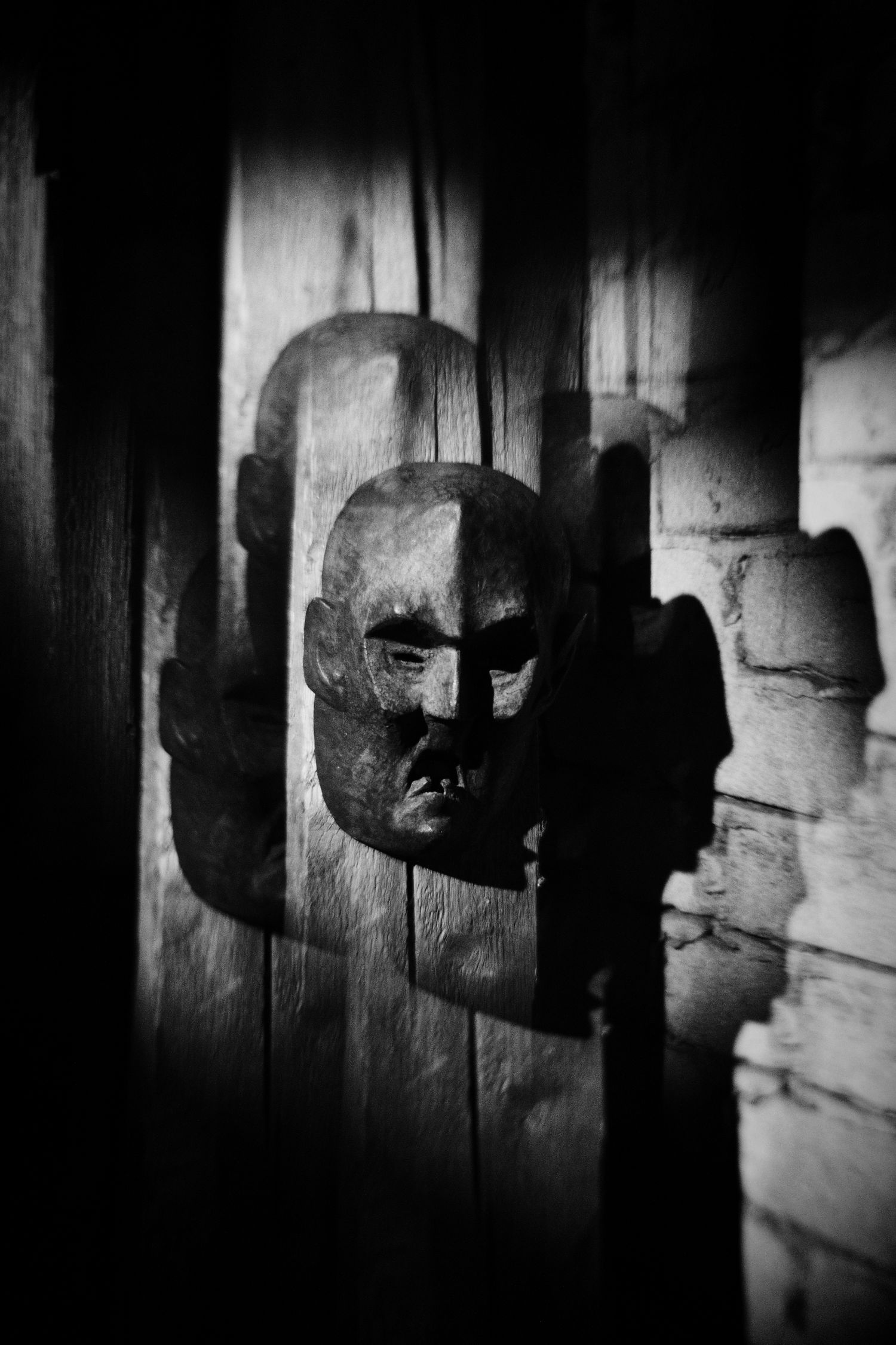 Black and white photo of a carved wooden face mask mounted on weathered wooden planks with dramatic shadows.