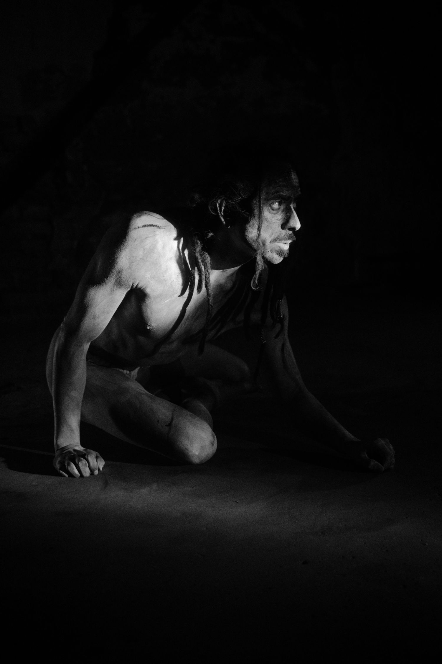 Black and white photo of a person crouching in darkness, body painted, in a dramatic theatrical pose.