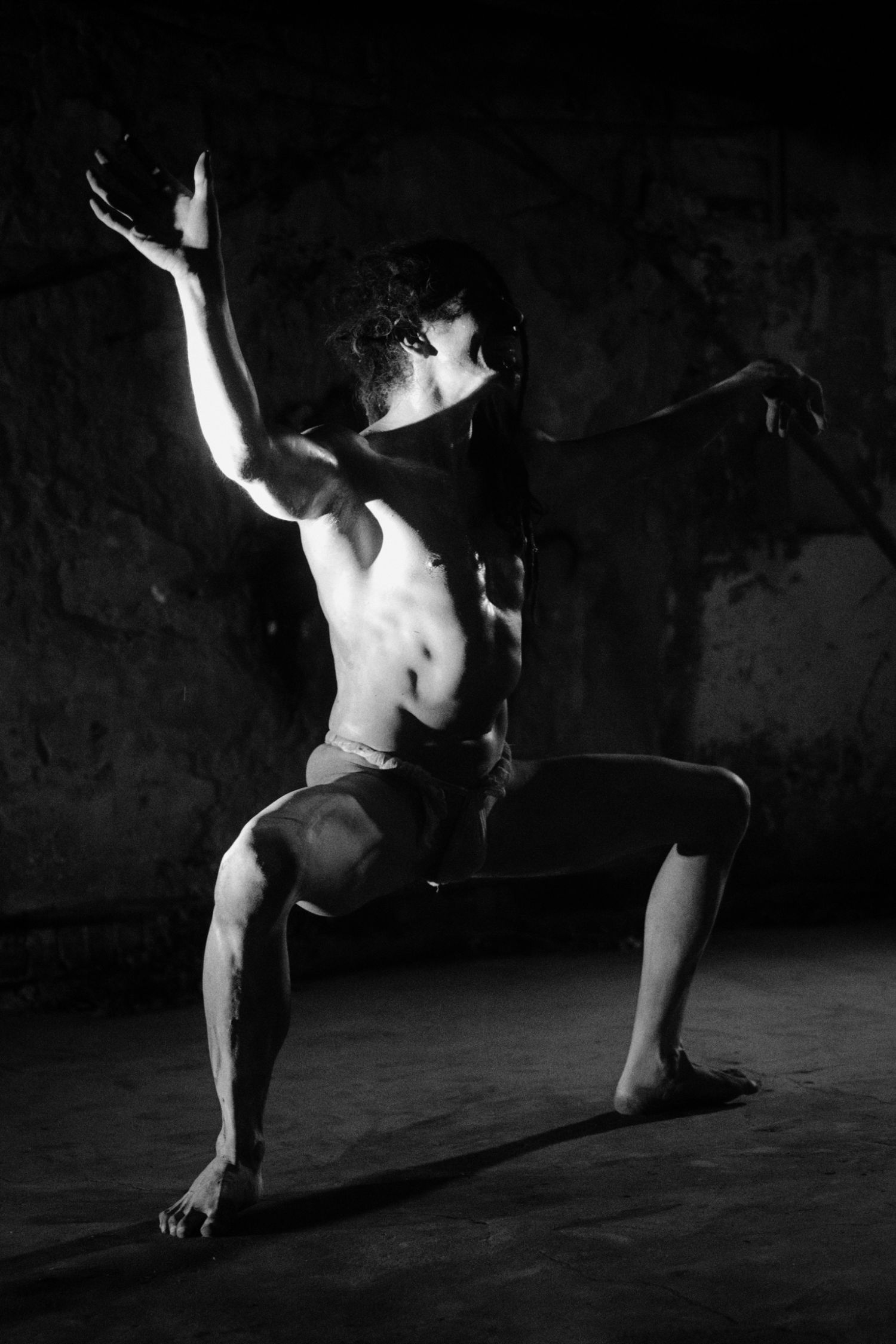 Black and white photo of a muscular male dancer in a dramatic lunge pose with one arm raised in a dark studio.