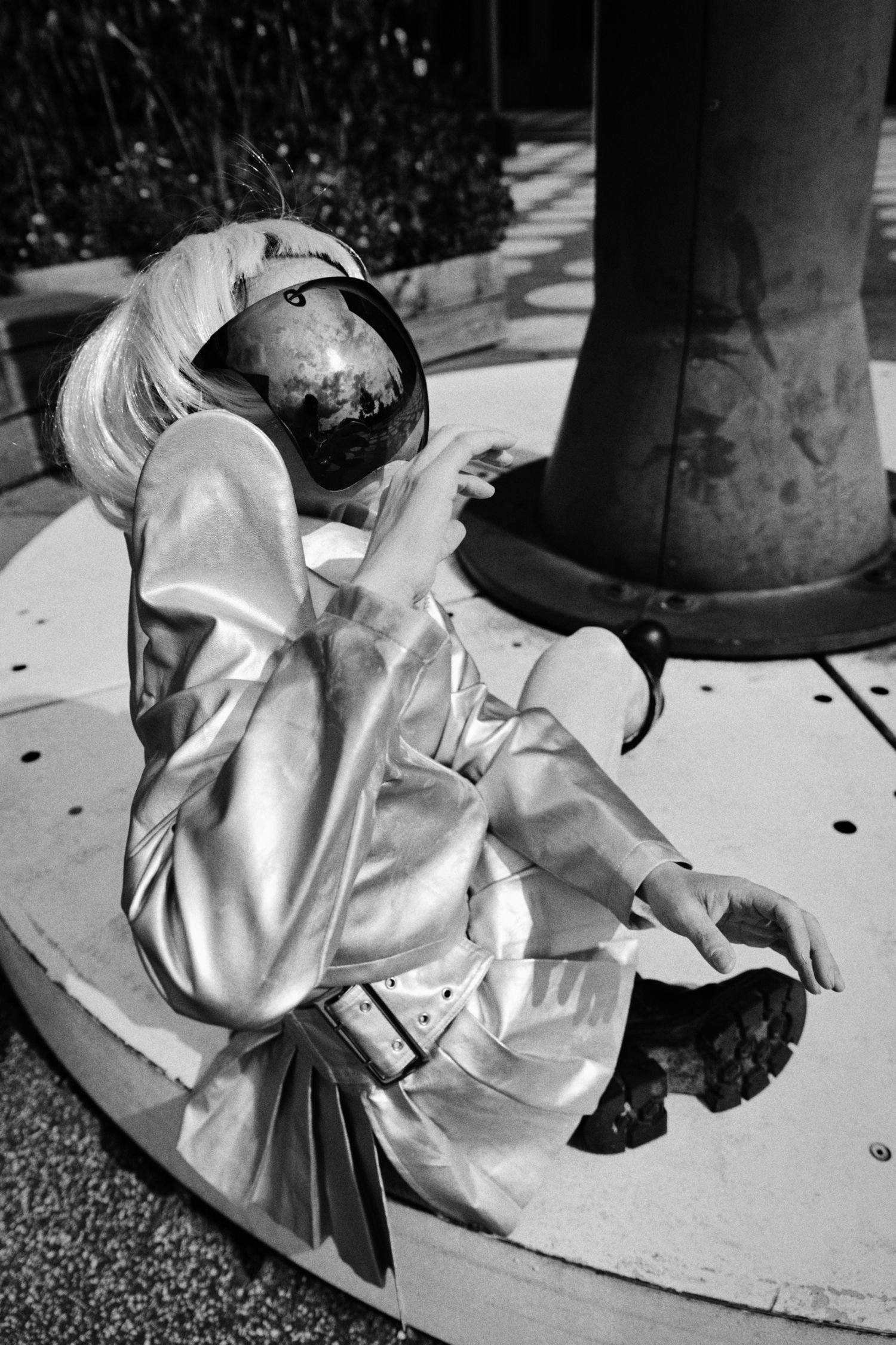 Black and white photo of a person in a shiny metallic trench coat and reflective space helmet sitting on a circular platform.